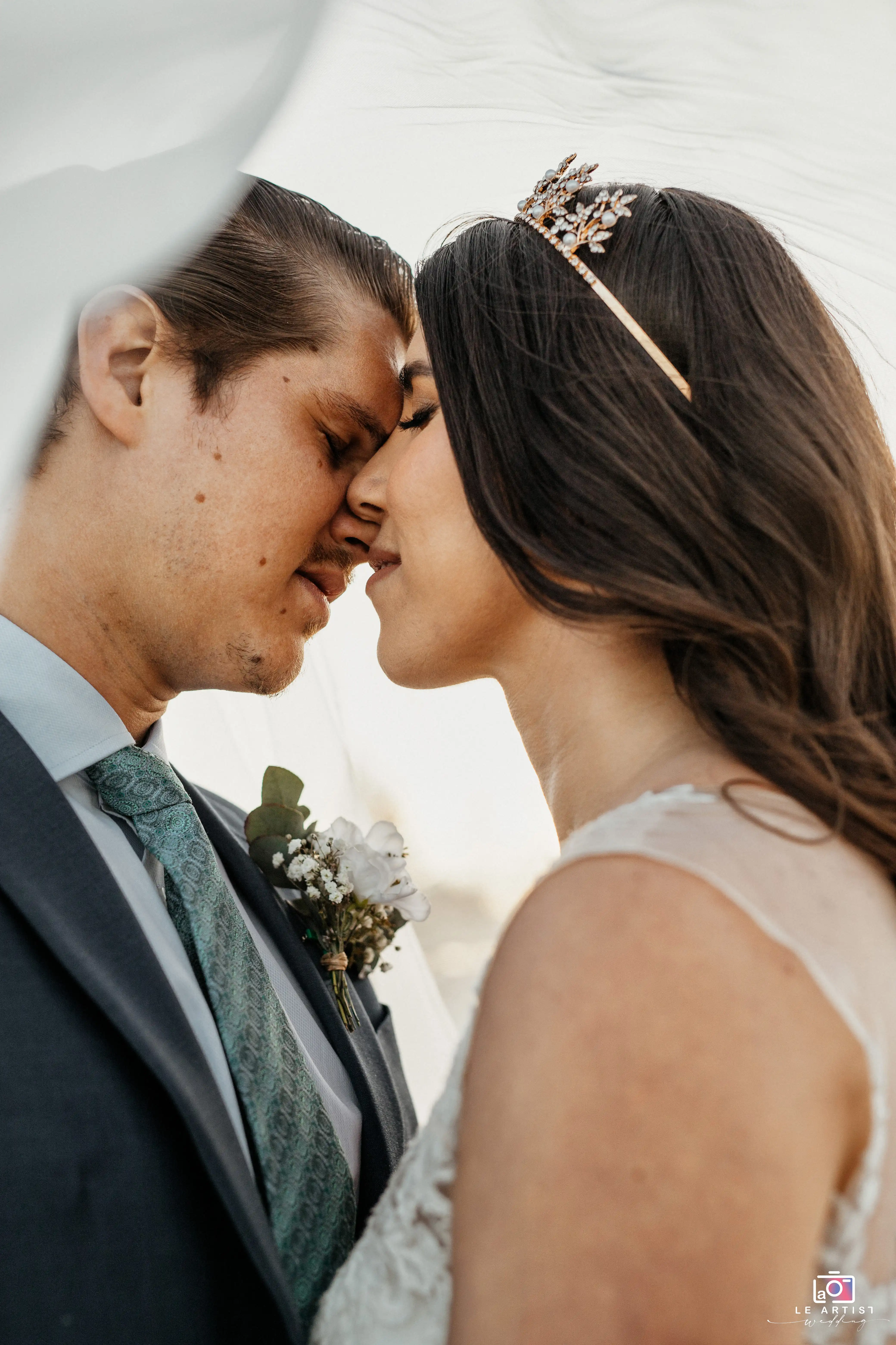 A tender moment captured as the couple leans in for a close embrace, highlighted by the bride's elegant headpiece and the groom's floral boutonniere. Le Artist Wedding