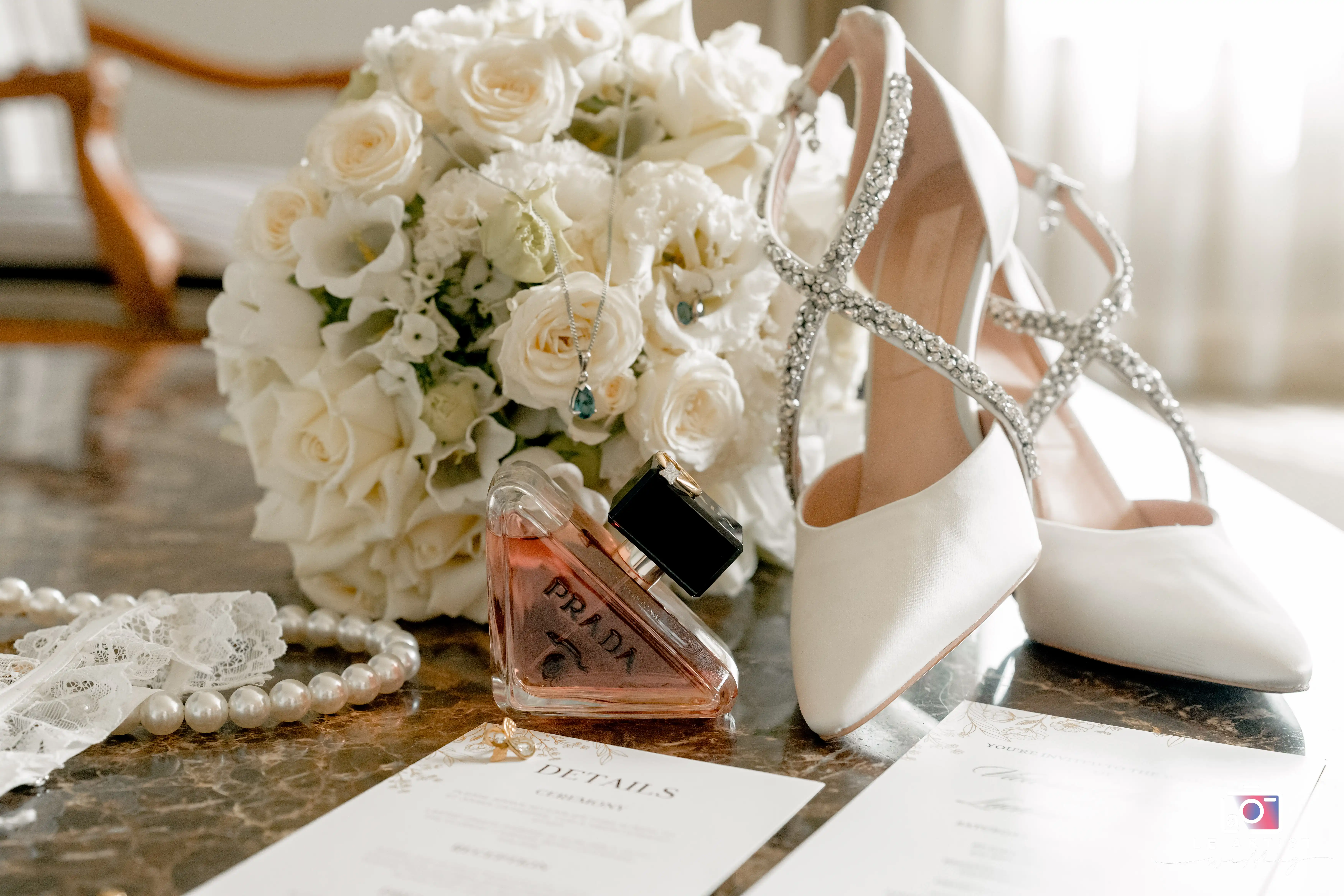 A beautifully arranged bridal detail shot featuring elegant white heels adorned with crystals, a lush bouquet of white roses, a delicate lace garter, a pearl necklace, and a sophisticated perfume bottle. The arrangement is set on a polished surface alongside wedding invitations, capturing the essence of the bride's style and elegance. Le Artist Wedding