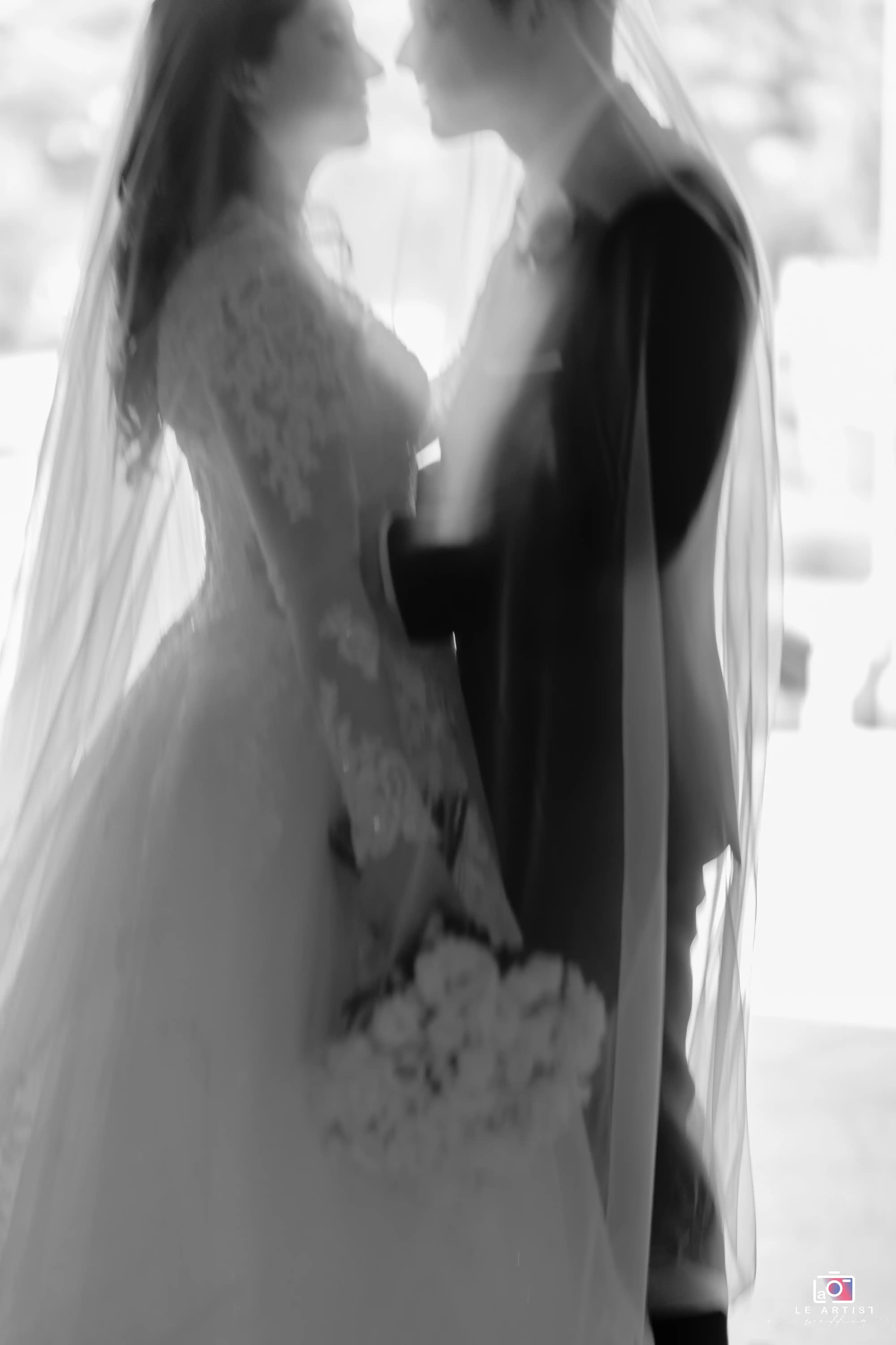 A dreamy black and white photograph capturing the tender moment of a bride and groom enveloped in each other's presence, the bride holds her bouquet while wearing an elegant lace gown. Le Artist Wedding