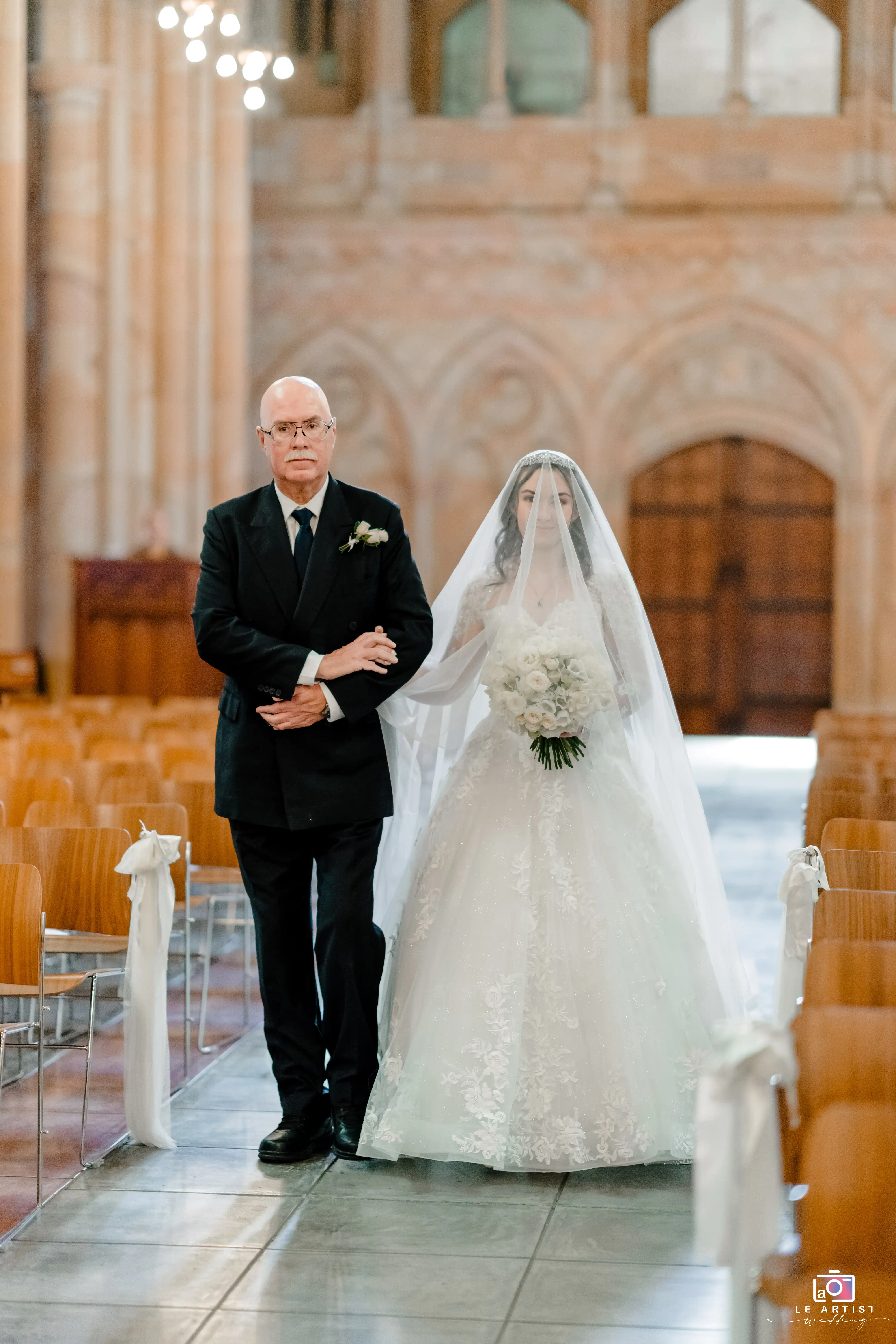 A bride is elegantly escorted down the aisle by her father, holding a bouquet of white flowers. Her veil delicately covers her face, while the beautiful church setting enhances the moment's solemnity. Le Artist Wedding