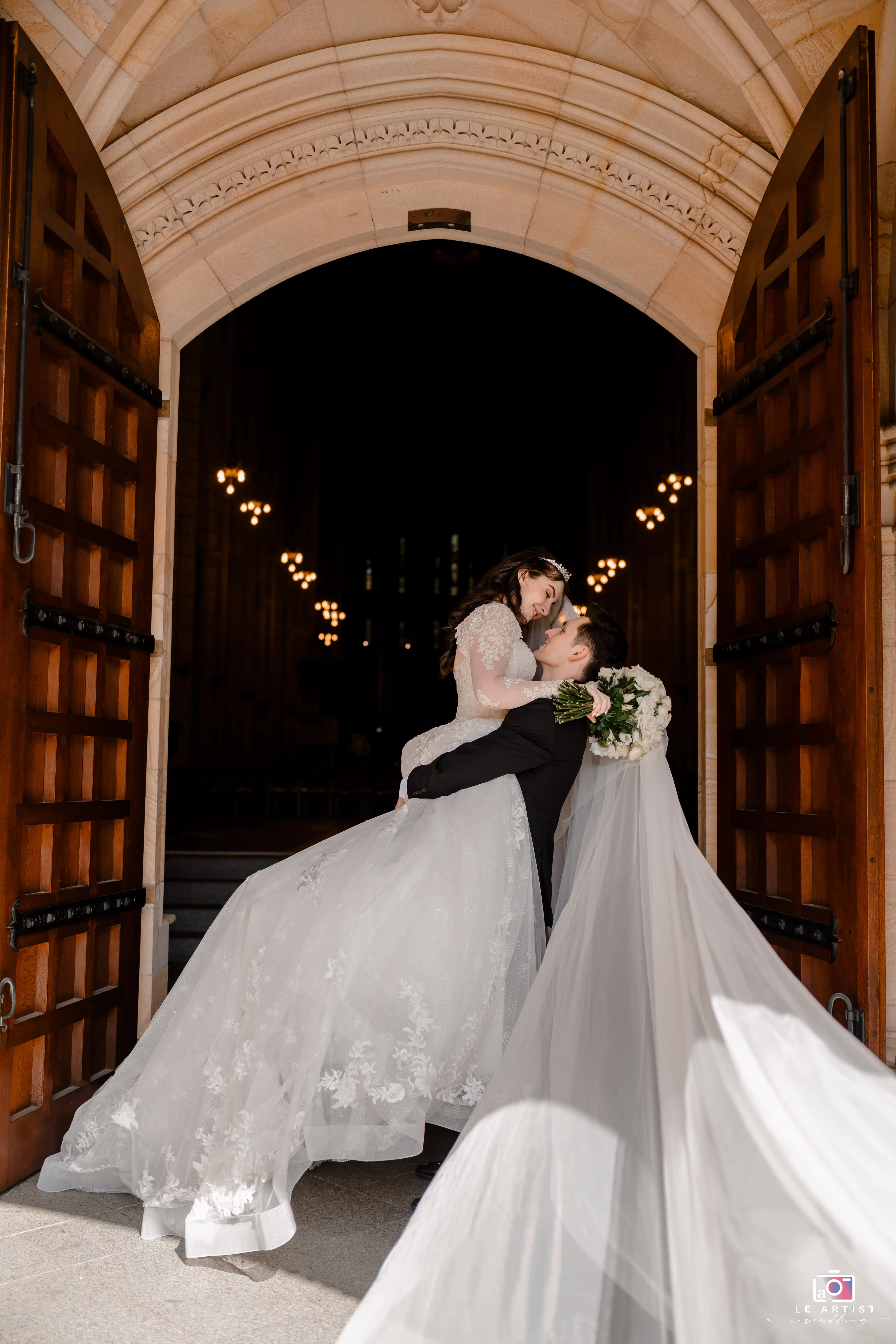 A romantic moment captures the groom sweeping his bride off her feet at the grand entrance of the venue. Le Artist Wedding