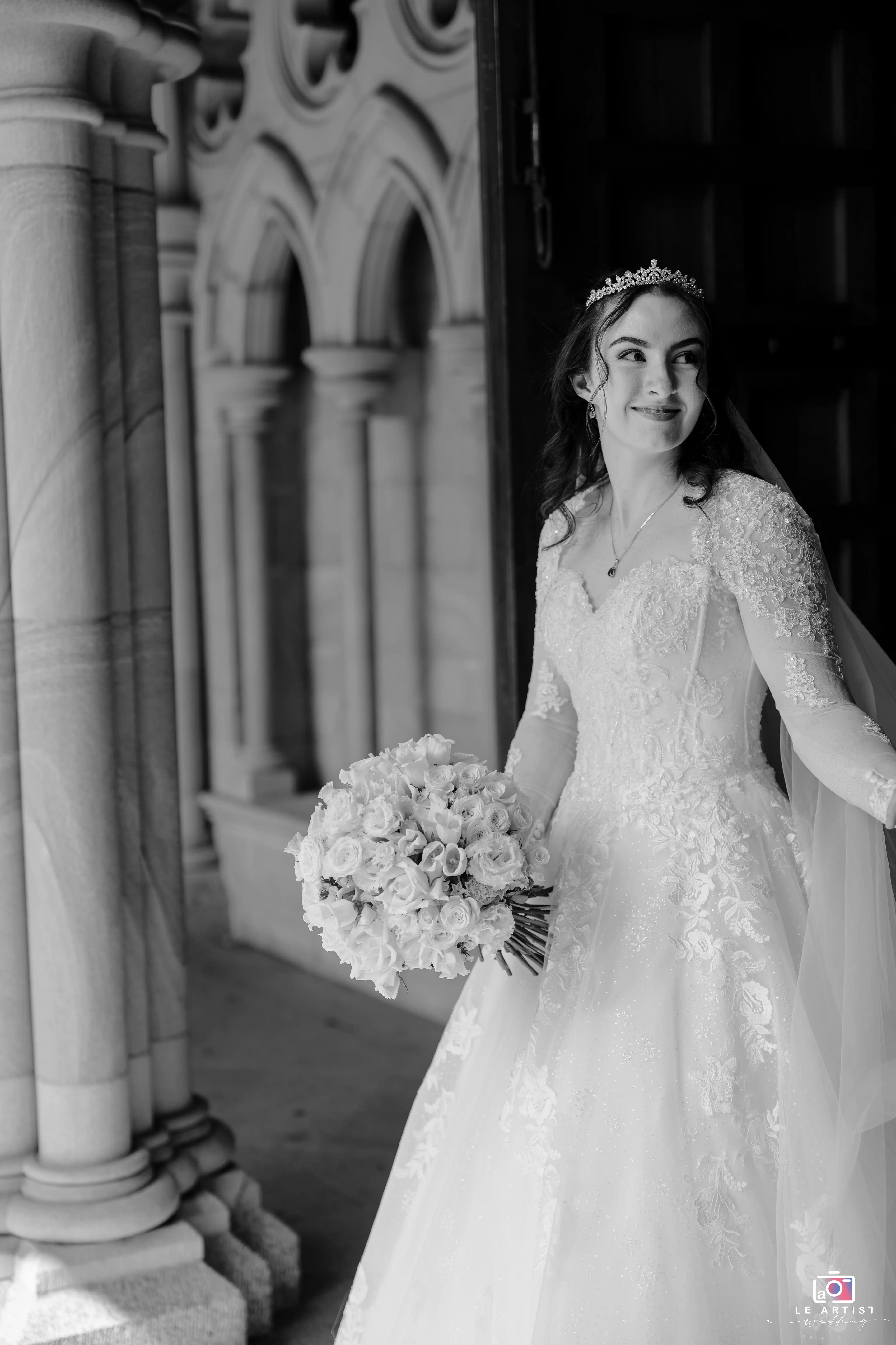 A bride radiantly smiles as she steps through an elegant archway, holding a bouquet of roses. Her gown's intricate embroidery adds to the timeless charm captured in this monochrome portrait. Le Artist Wedding