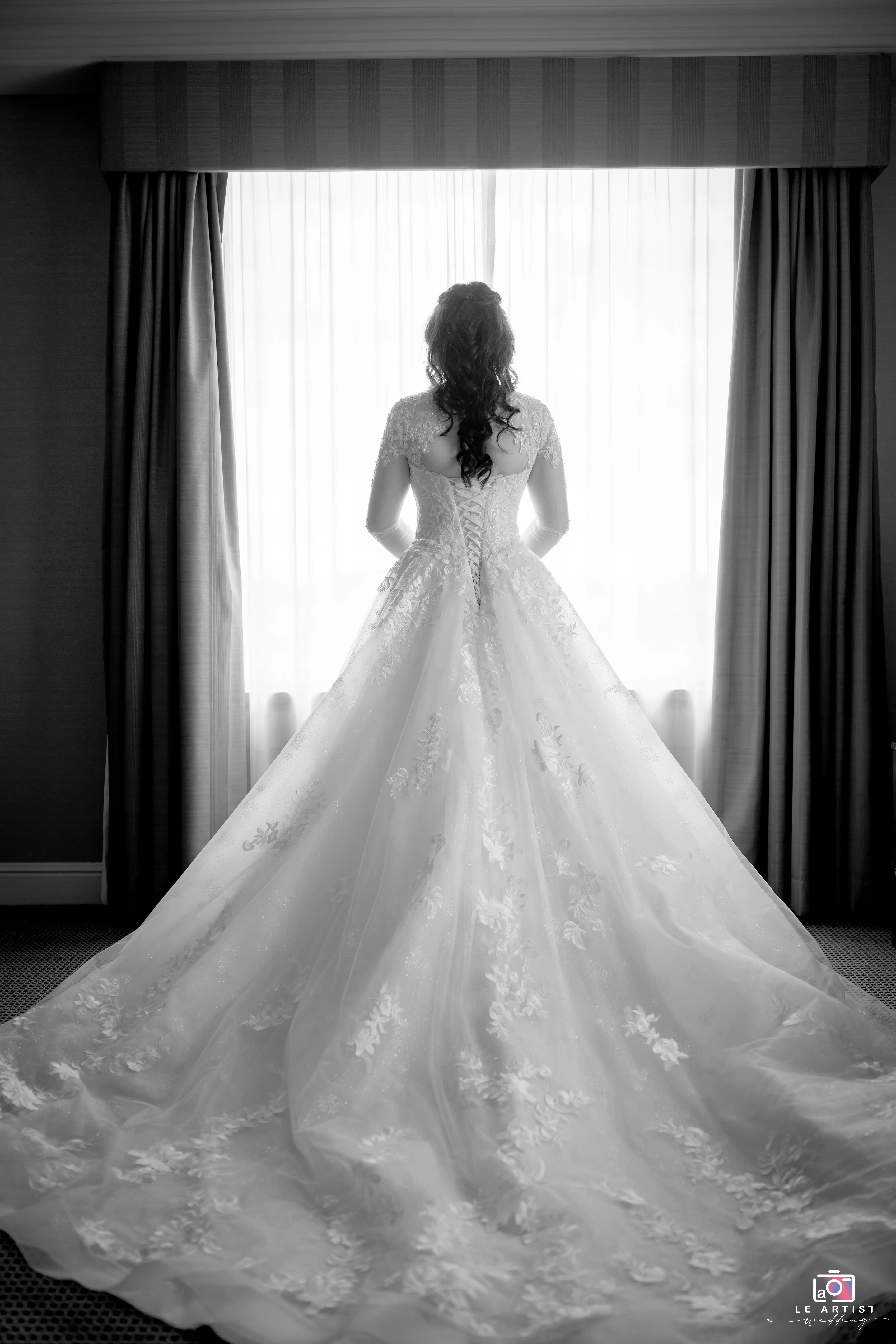 A stunning back pose of the bride showcasing her elegant lace wedding gown in front of a softly lit window. Le Artist Wedding