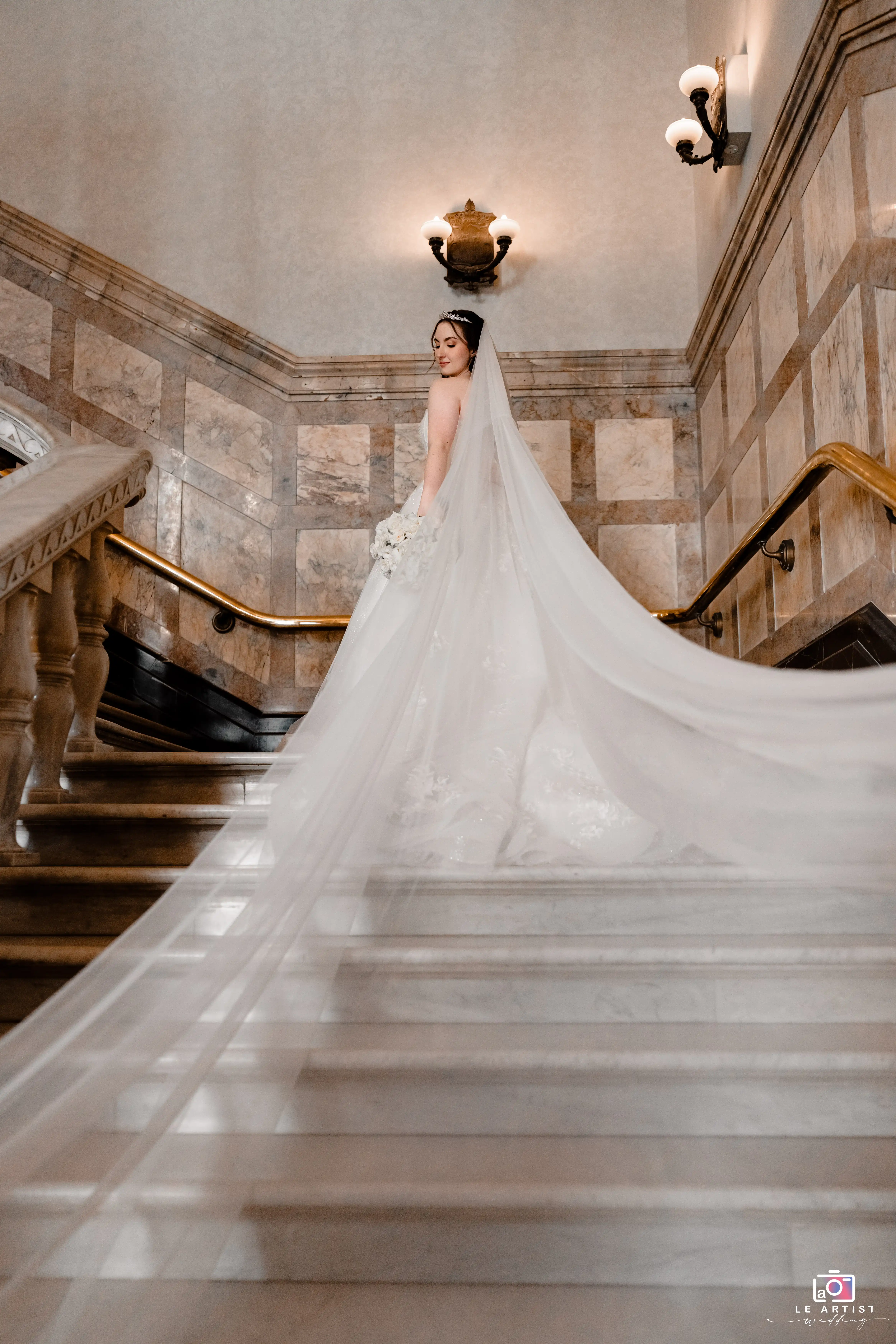 The bride gracefully ascends a grand staircase, her long veil trailing elegantly behind her, creating a timeless and majestic scene. Le Artist Wedding