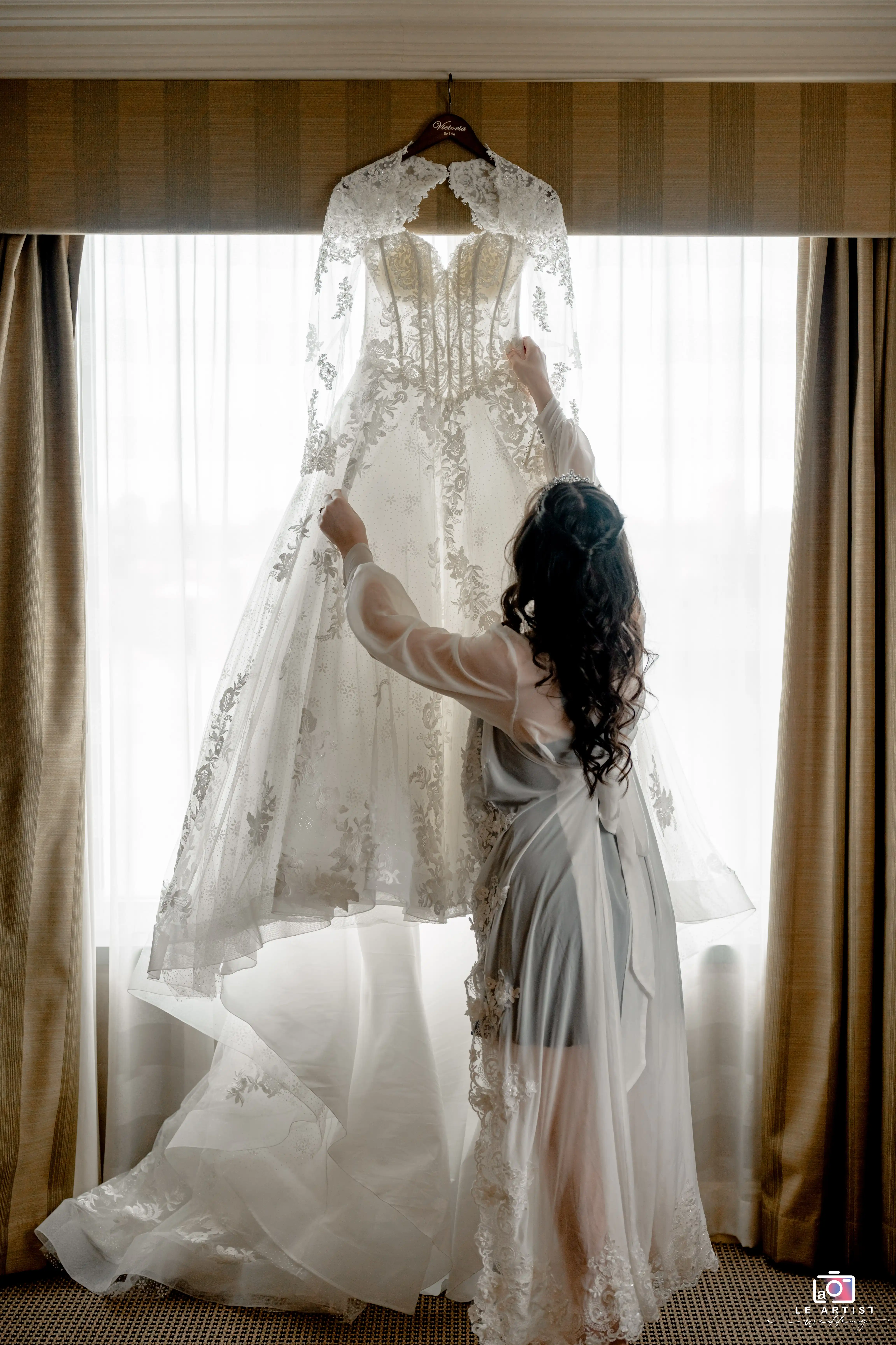 A bride admires her intricately detailed gown hanging by the window, capturing a moment of anticipation before the ceremony. Le Artist Wedding