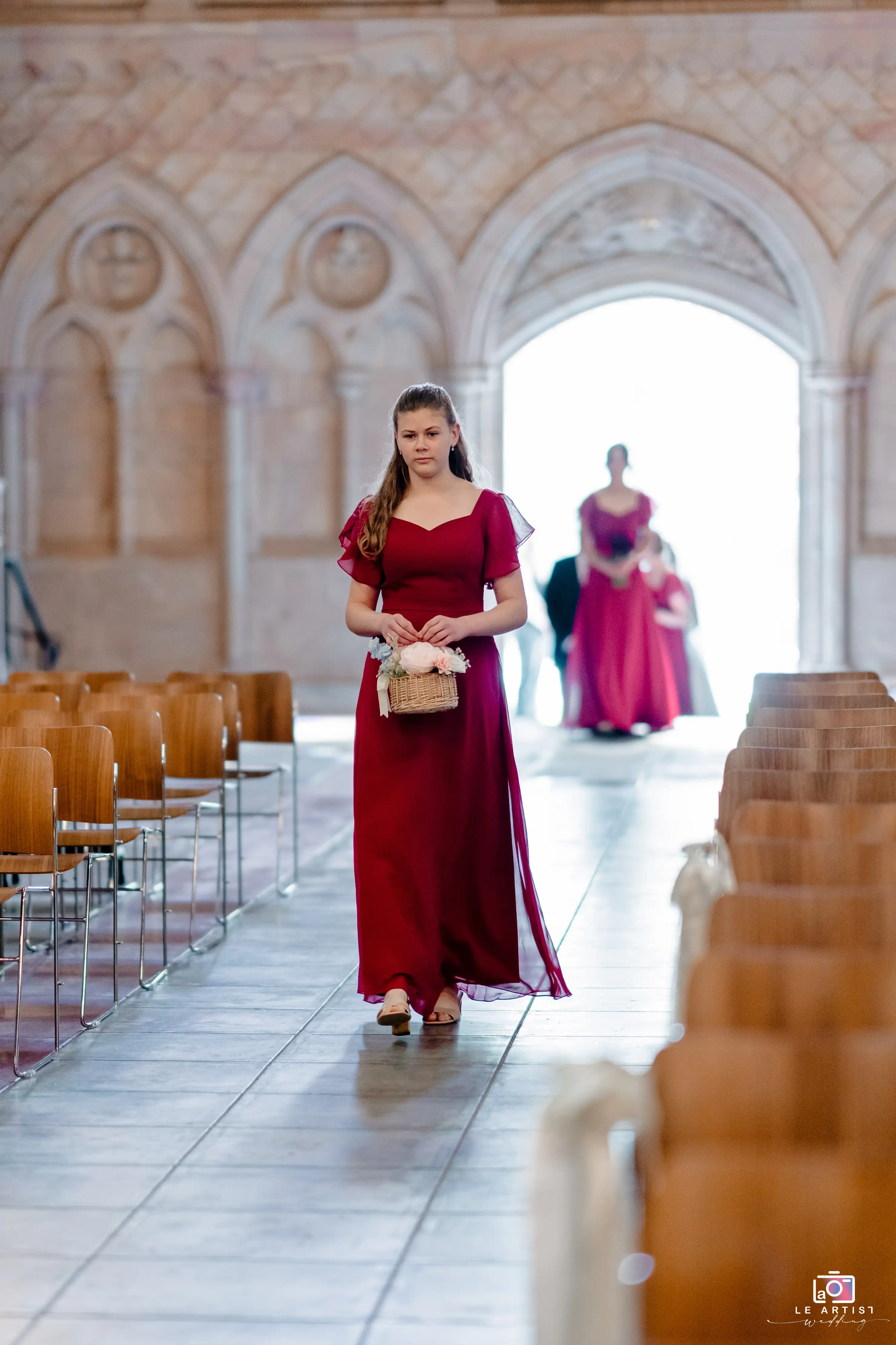 A bridesmaid in an elegant burgundy dress walks down the aisle in a beautifully decorated church, holding a basket of flowers. Le Artist Wedding