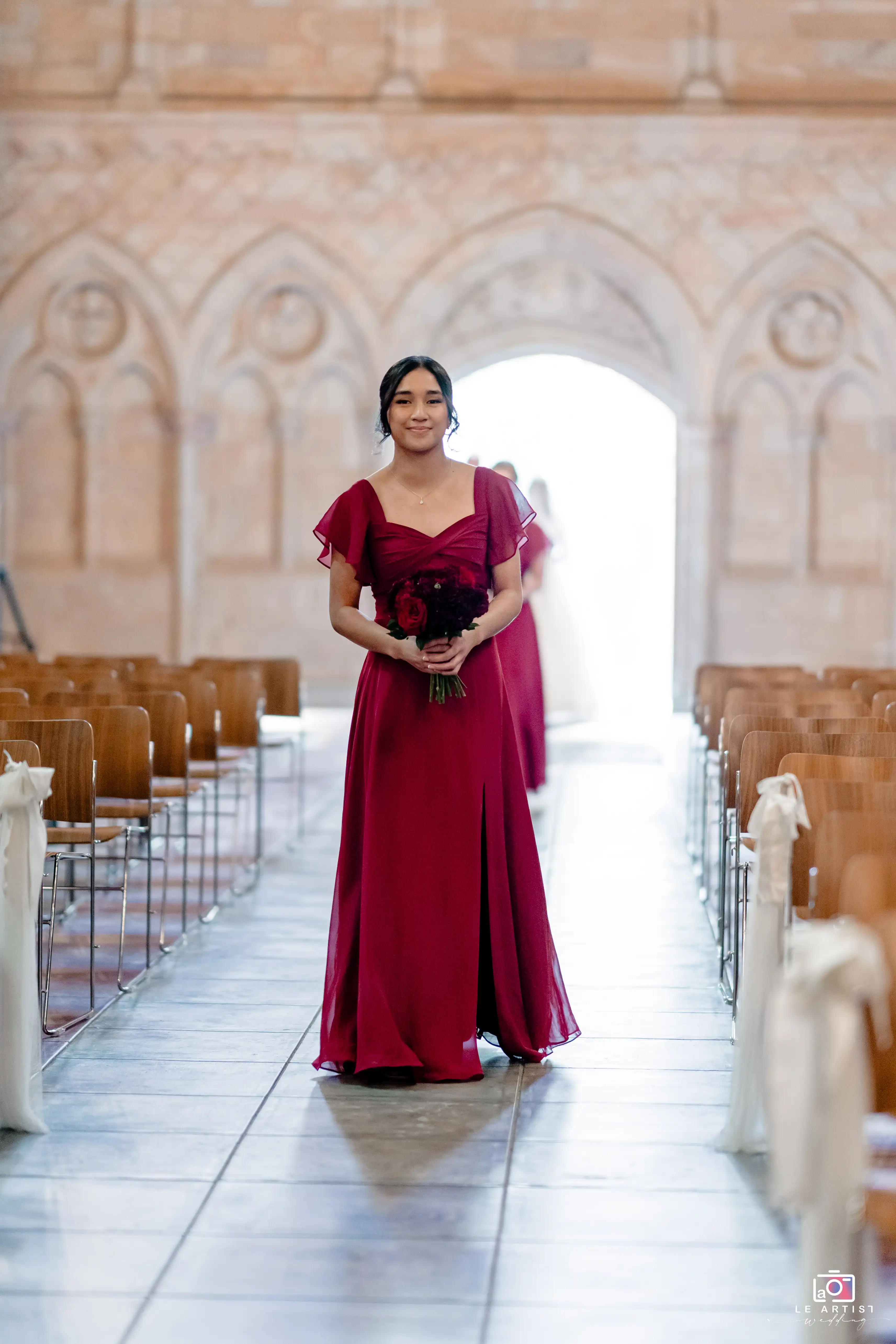 A bridesmaid elegantly walks down the aisle in a deep red dress, carrying a bouquet of matching flowers, in a beautifully adorned venue. Le Artist Wedding