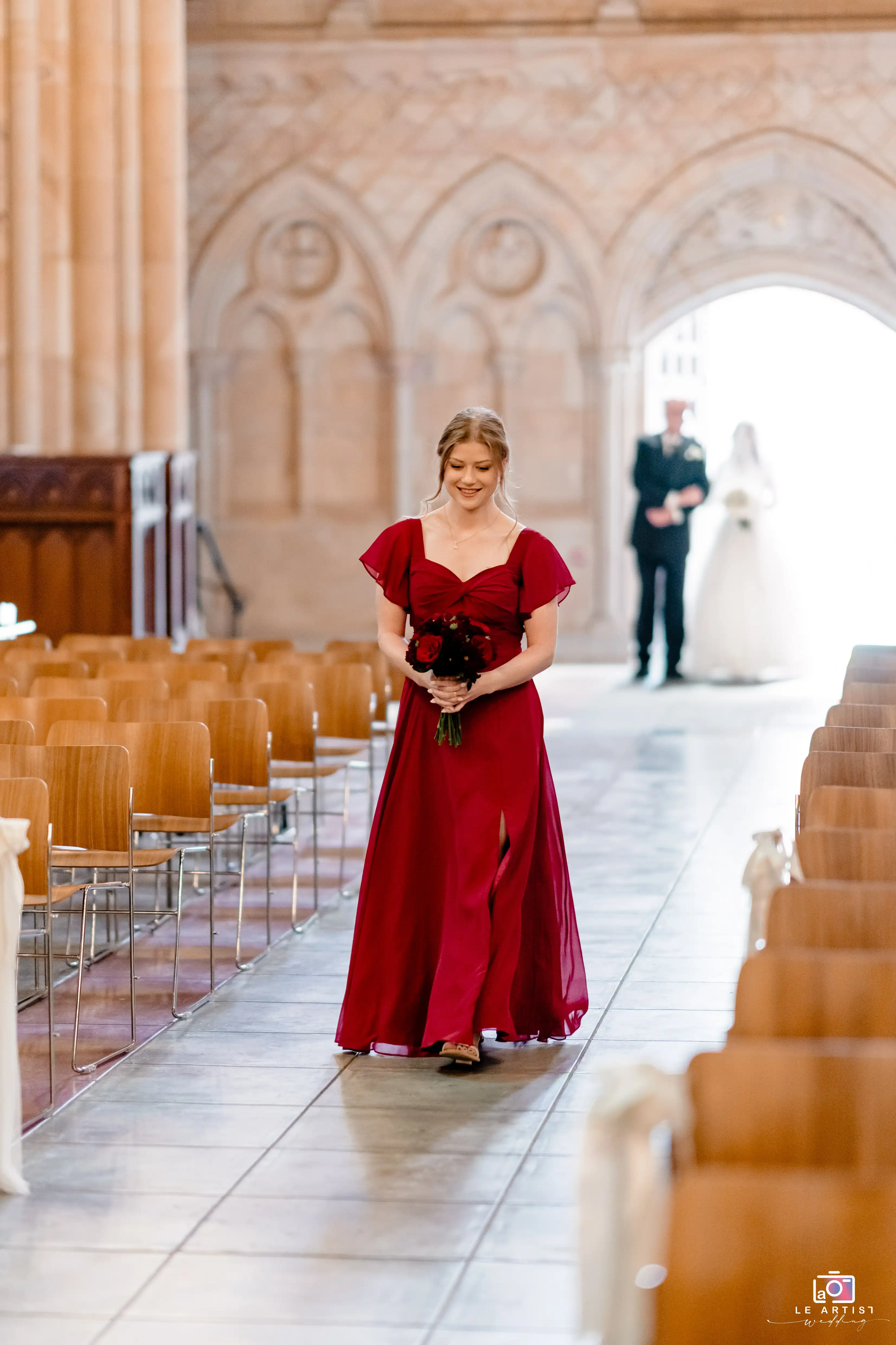 A bridesmaid in a striking red dress walks gracefully down the aisle, holding a bouquet, with the bride and groom visible in the background. The scene is set in an elegantly decorated church. Le Artist Wedding