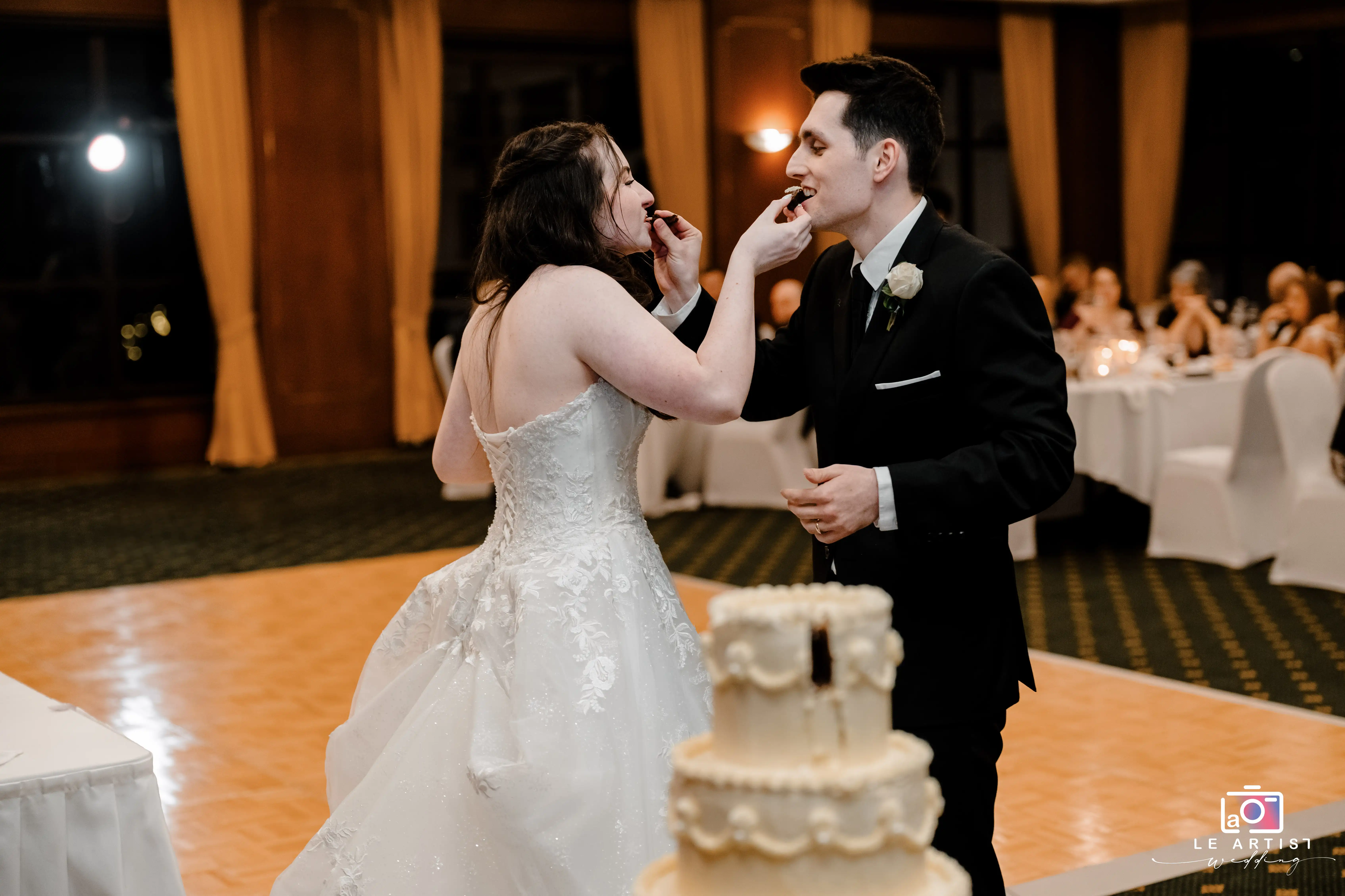 The couple shares a playful moment feeding each other cake, capturing a joyful tradition during their wedding celebration. Le Artist Wedding