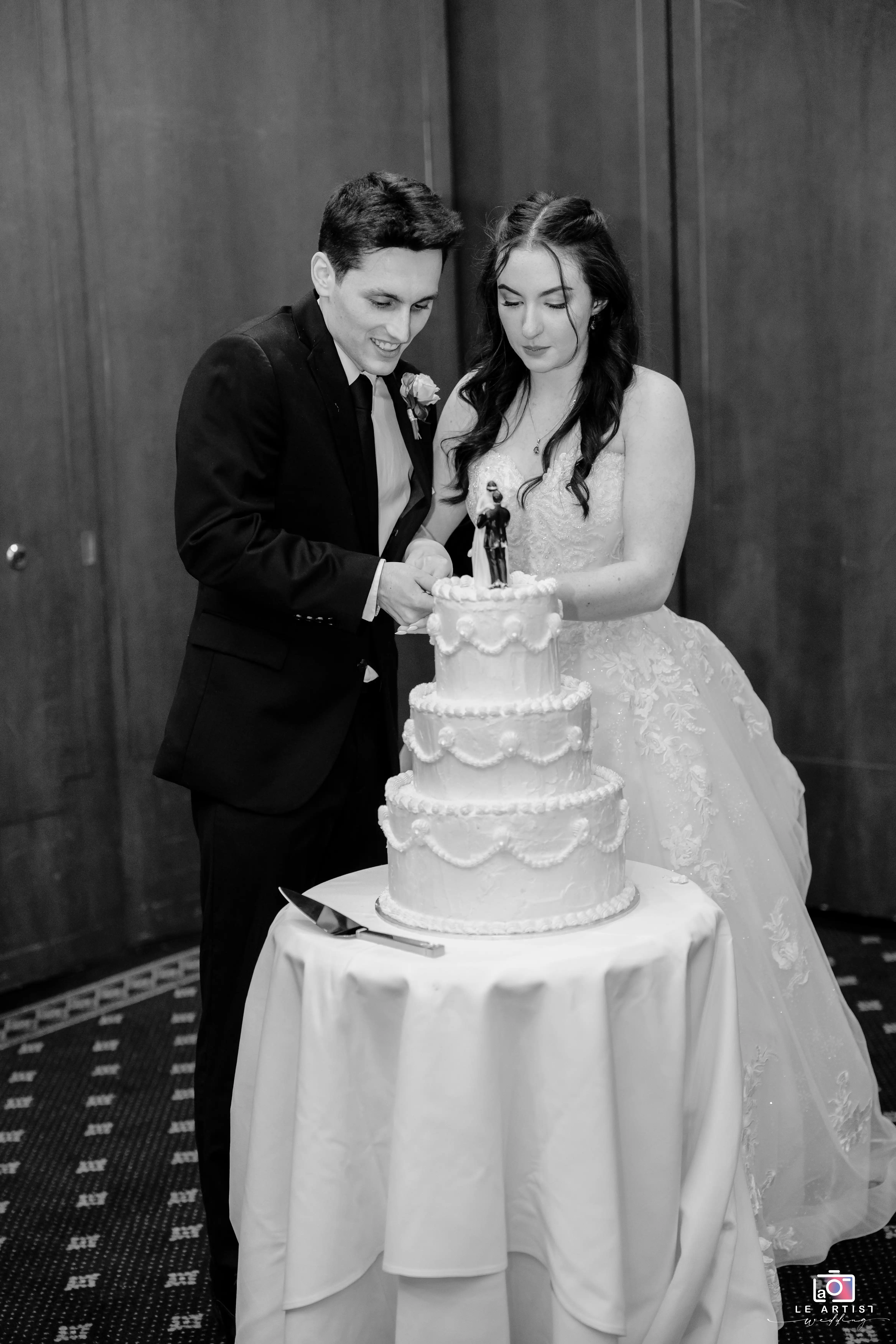 The couple shares a heartfelt moment as they cut their elegant tiered wedding cake together. Le Artist Wedding