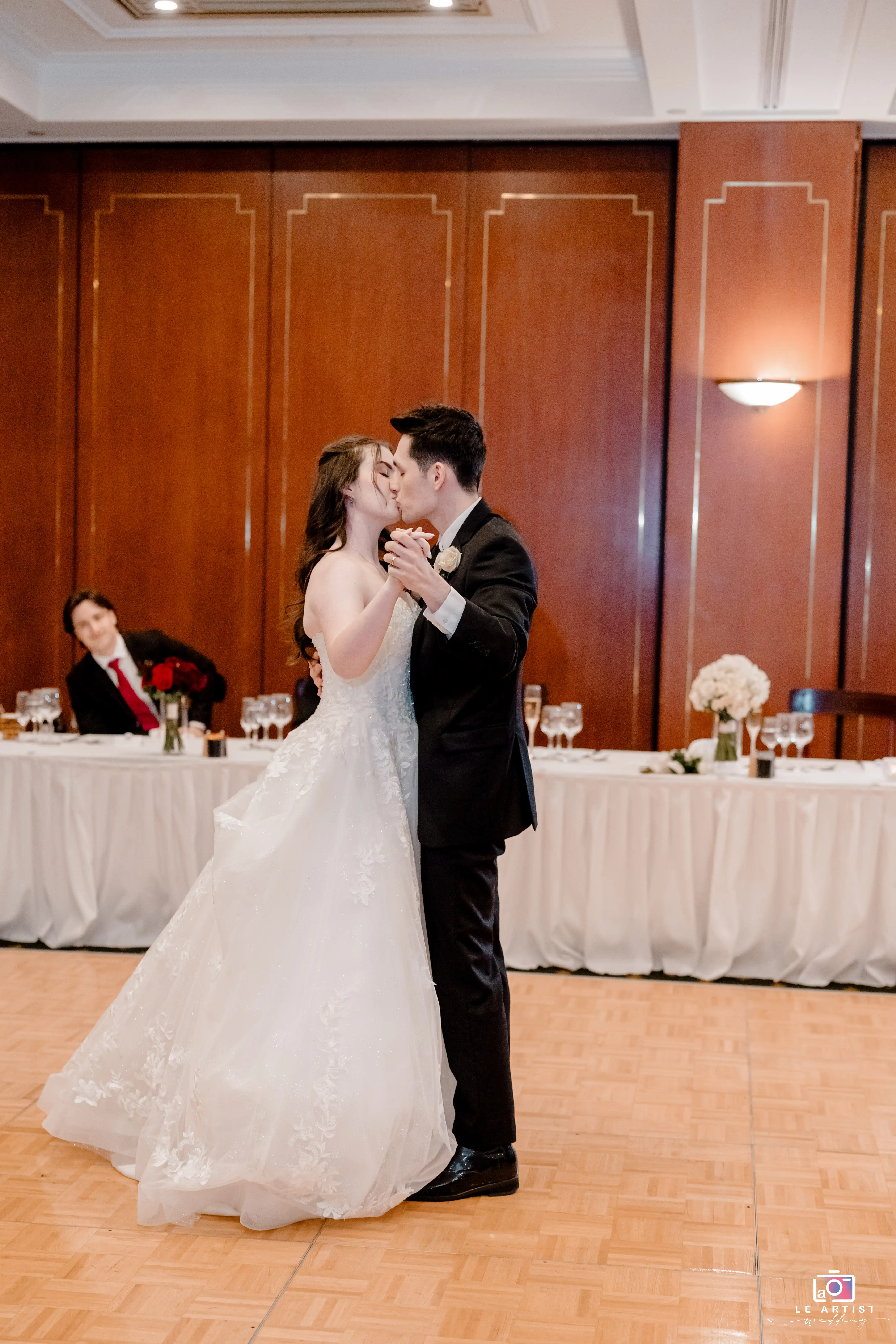 The couple shares a romantic kiss during their first dance at the wedding reception, with a beautifully set head table in the background. Le Artist Wedding