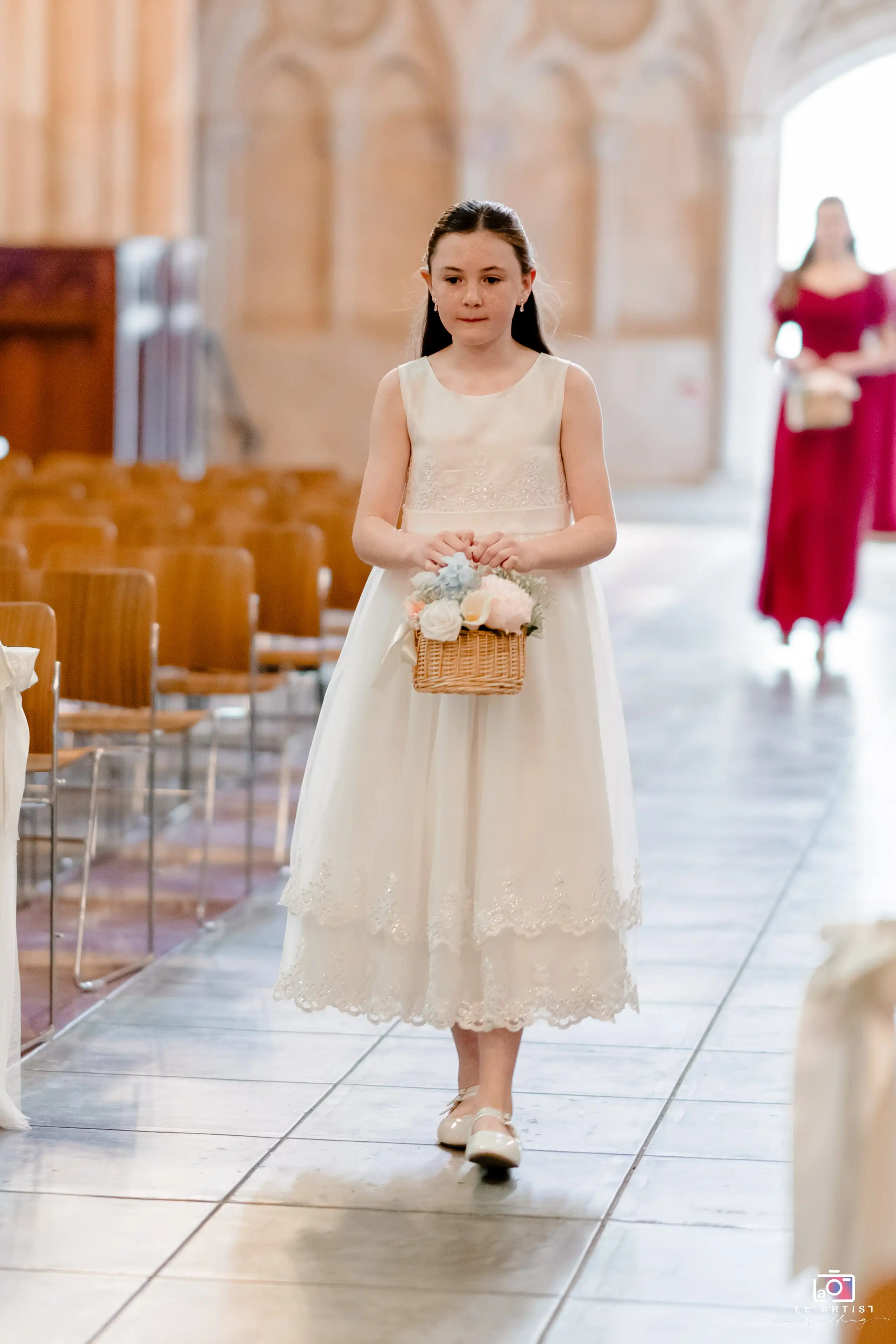 A young flower girl in an elegant white dress carries a basket of flowers as she walks down the aisle, setting a delicate and charming tone for the wedding ceremony. Le Artist Wedding