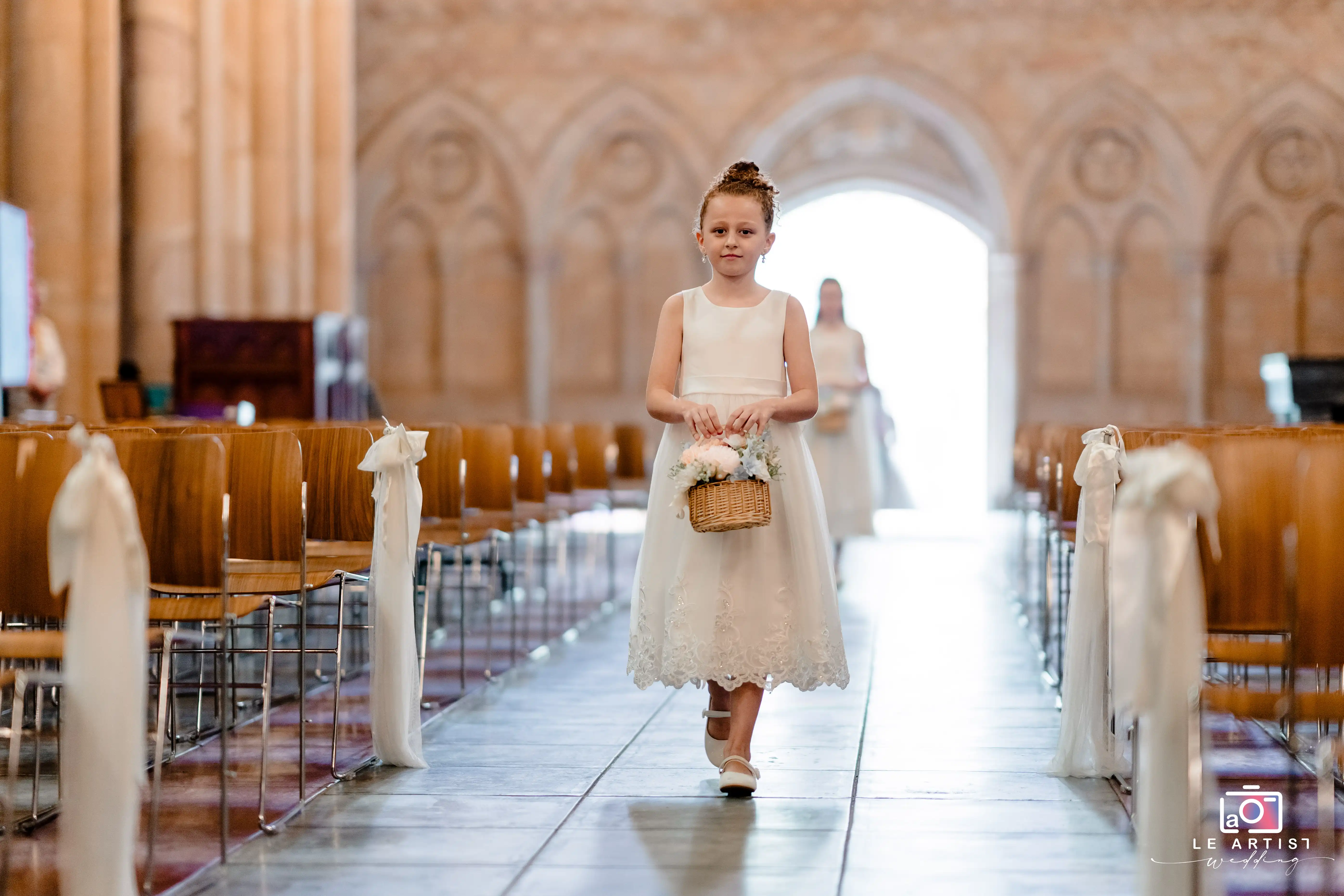 A young flower girl gracefully walks down the aisle, holding a basket of flowers, in a beautifully decorated church. Le Artist Wedding