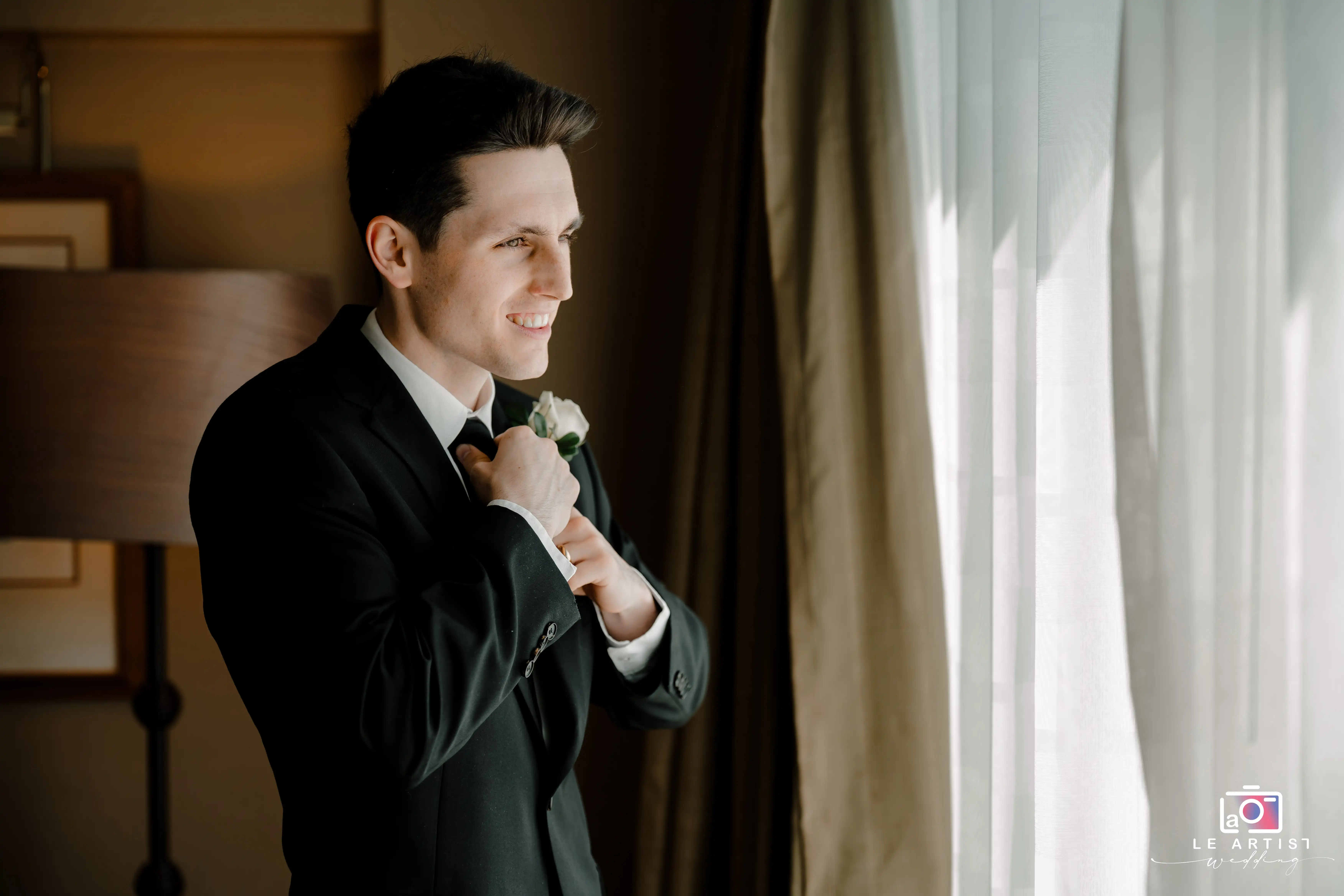 The groom prepares for his wedding day, fastening his tie and smiling with anticipation. Le Artist Wedding
