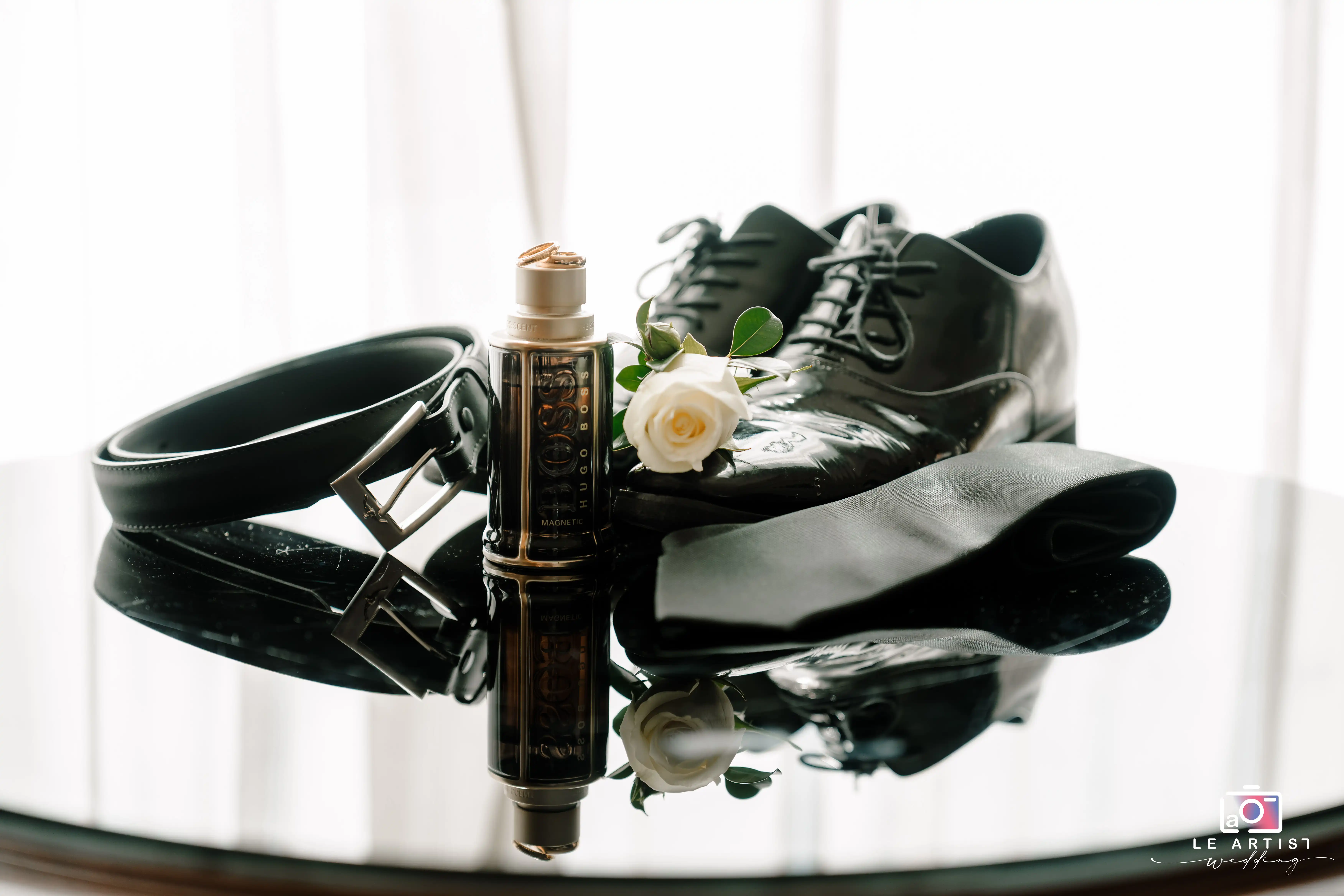 A stylish collection of groom's accessories, including polished shoes, a sleek belt, cologne, and a white rose boutonniere, arranged elegantly on a reflective surface. Le Artist Wedding