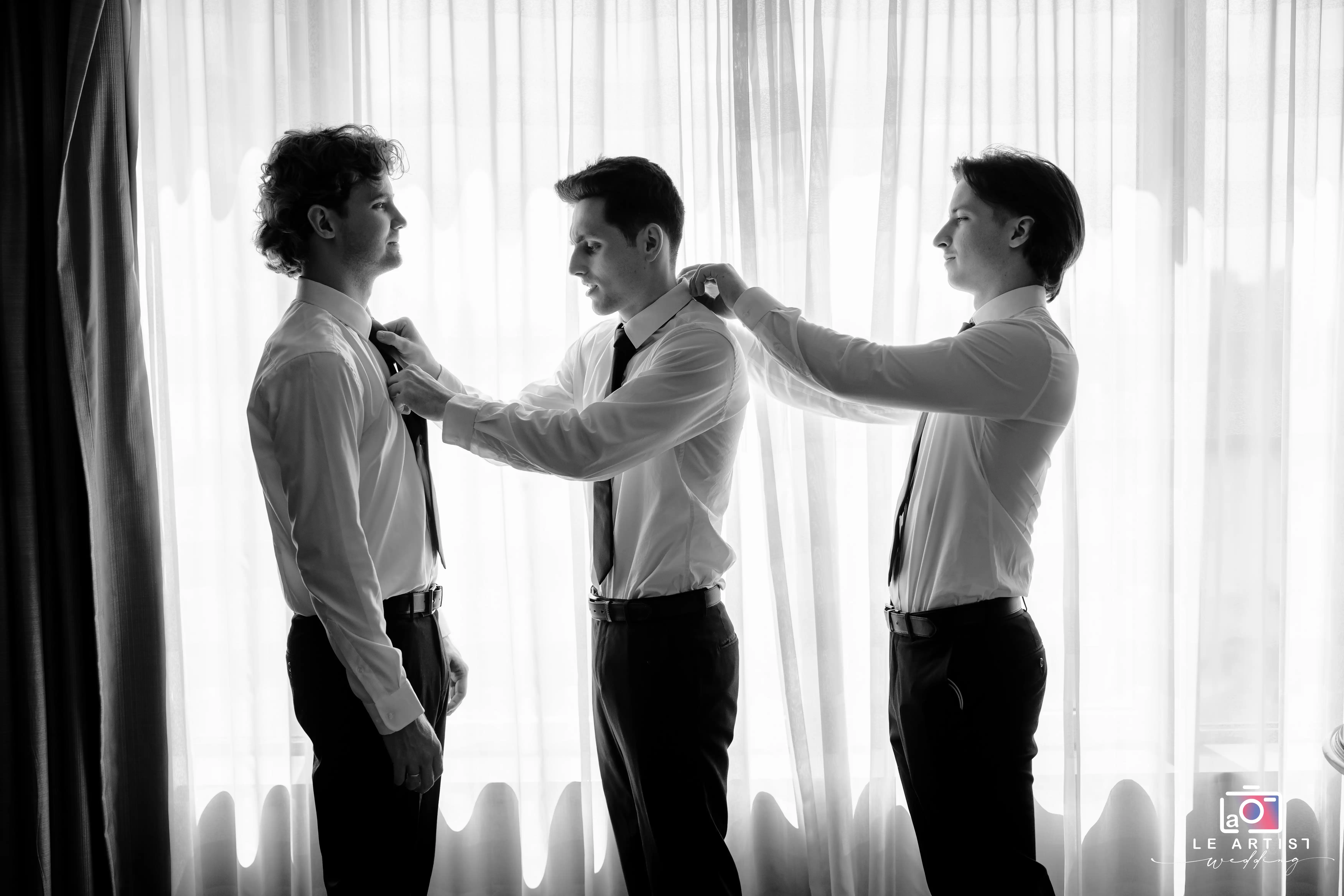 Groomsmen assist each other with their ties, preparing for the wedding ceremony in a candid moment by the window. Le Artist Wedding