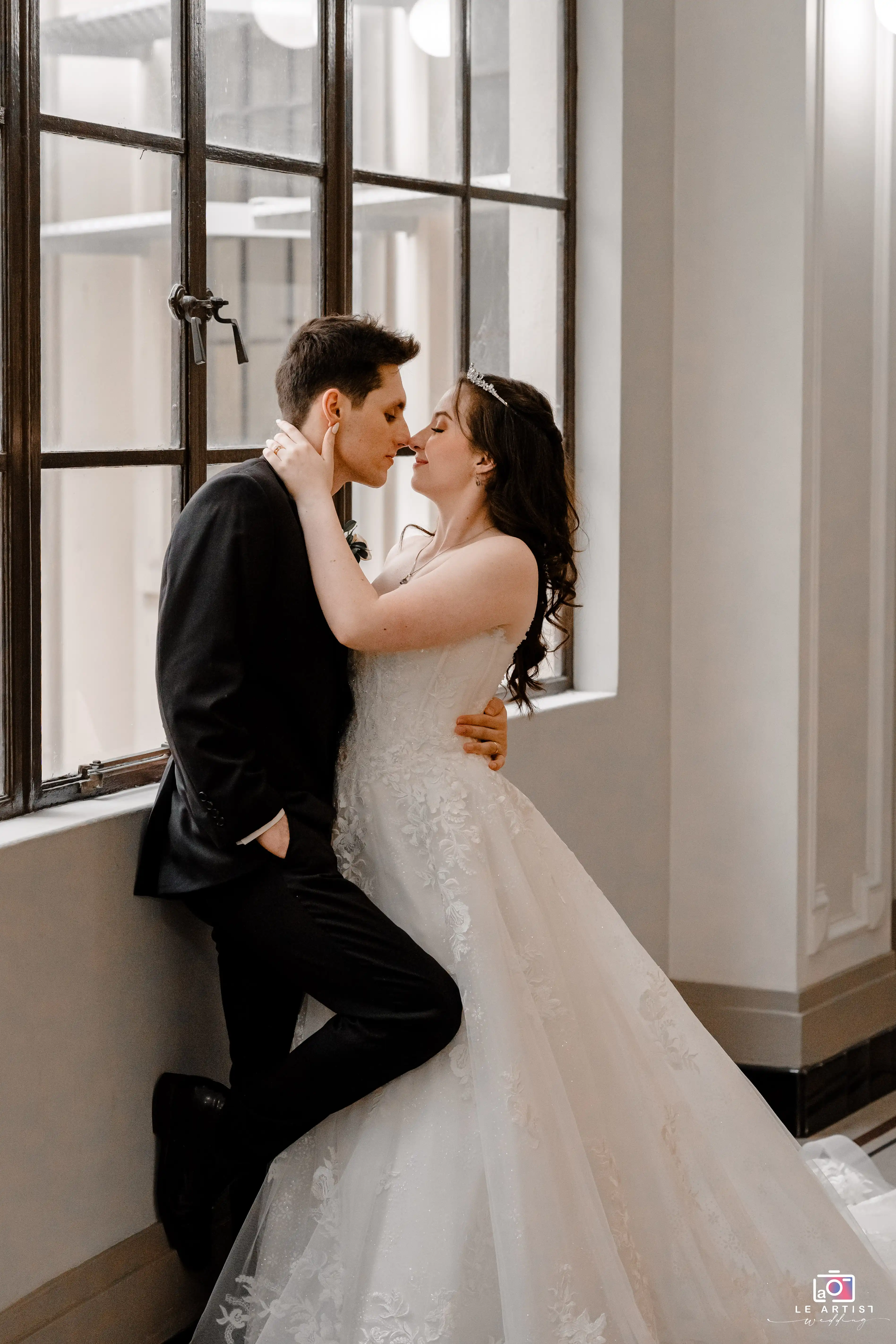 The couple shares an intimate moment by a sunlit window, capturing the tenderness of their wedding day. Le Artist Wedding
