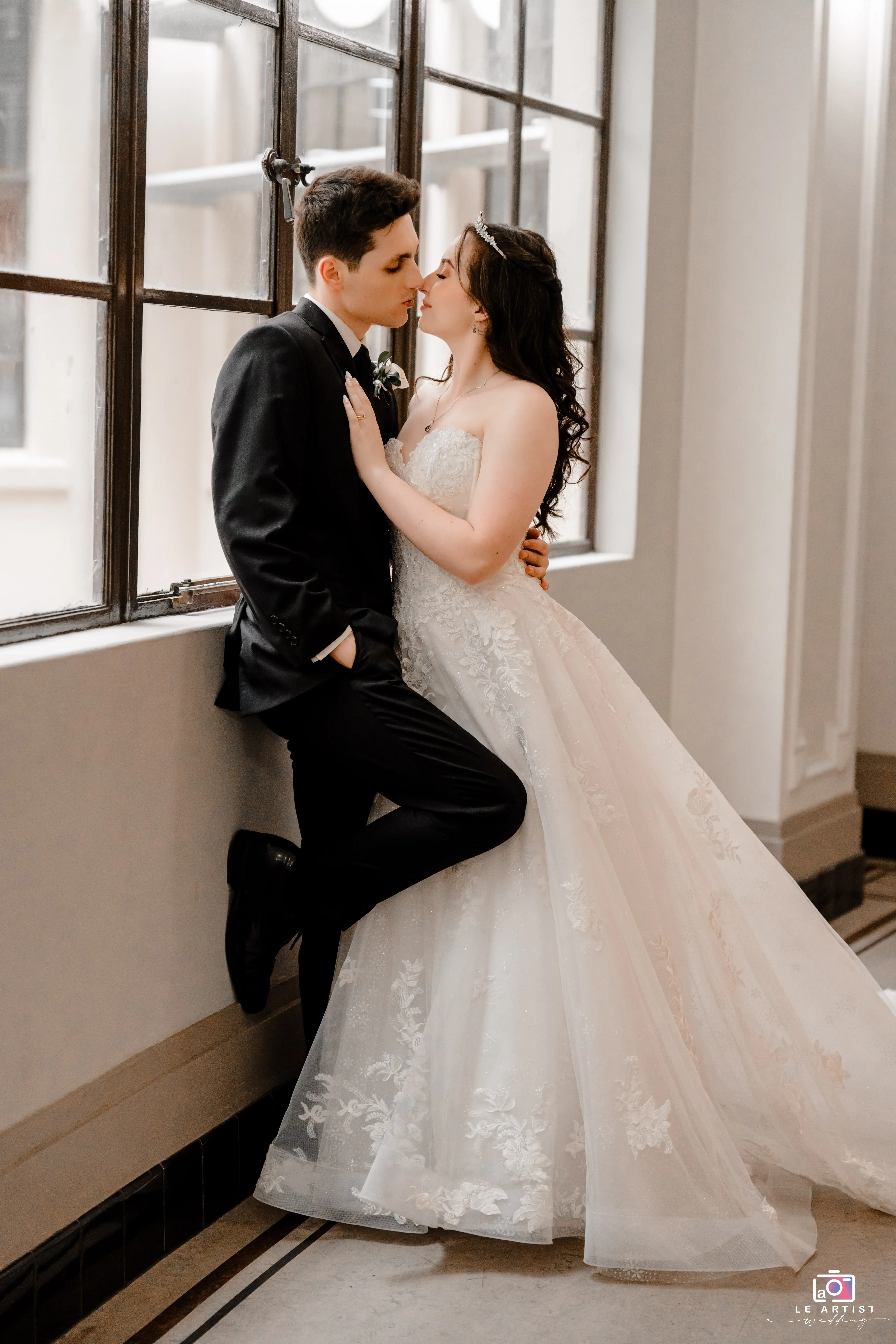 The couple shares an intimate moment by the window, capturing the essence of romance on their special day. Le Artist Wedding