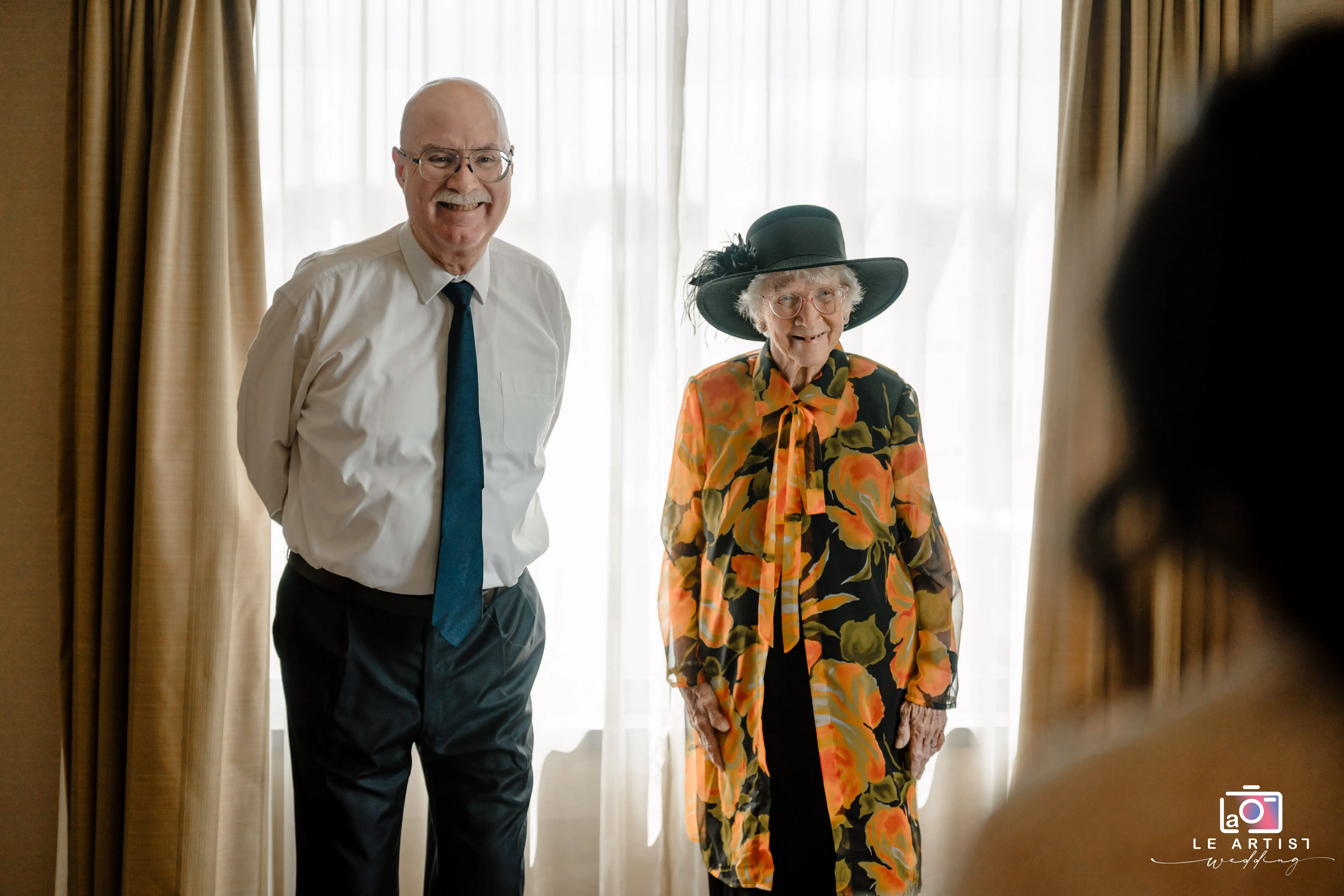A joyful moment between two guests captured with natural light streaming softly through the curtains. Le Artist Wedding