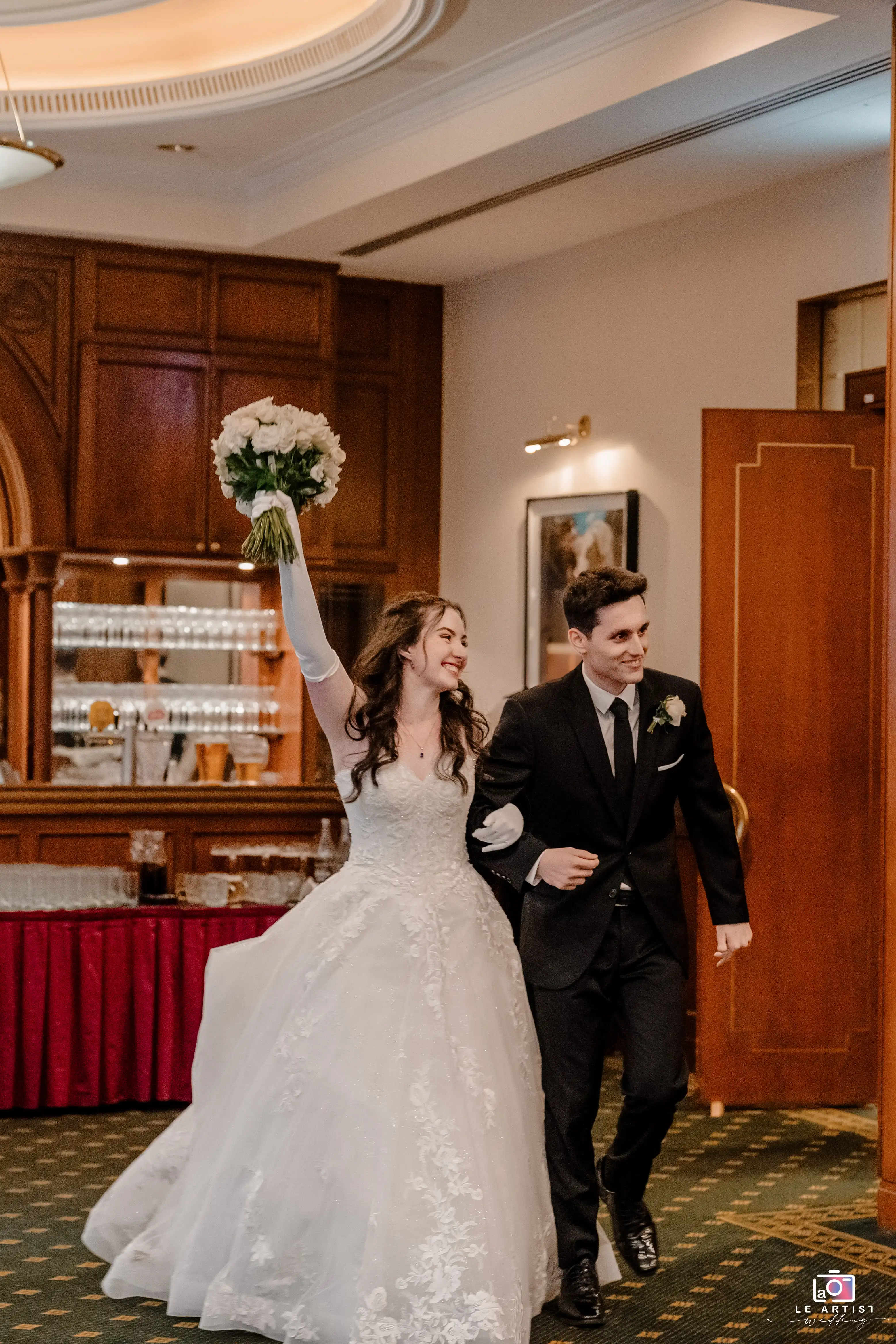 The newlyweds joyfully enter the reception, expressing their excitement and happiness with raised arms and bright smiles. Le Artist Wedding