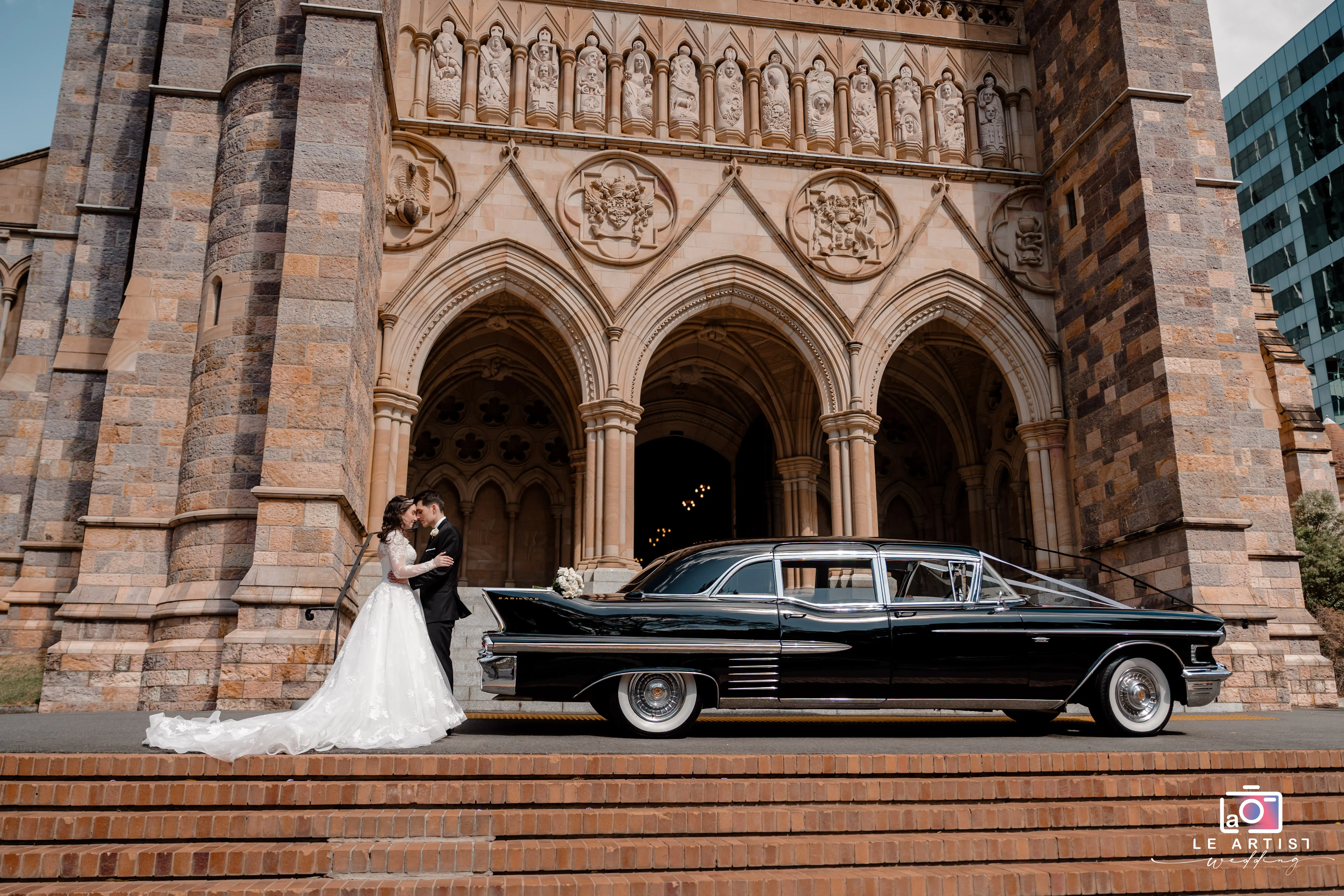 The newlyweds share an intimate moment on the steps of a magnificent stone cathedral, with a classic black car parked alongside, adding a touch of vintage elegance. Le Artist Wedding