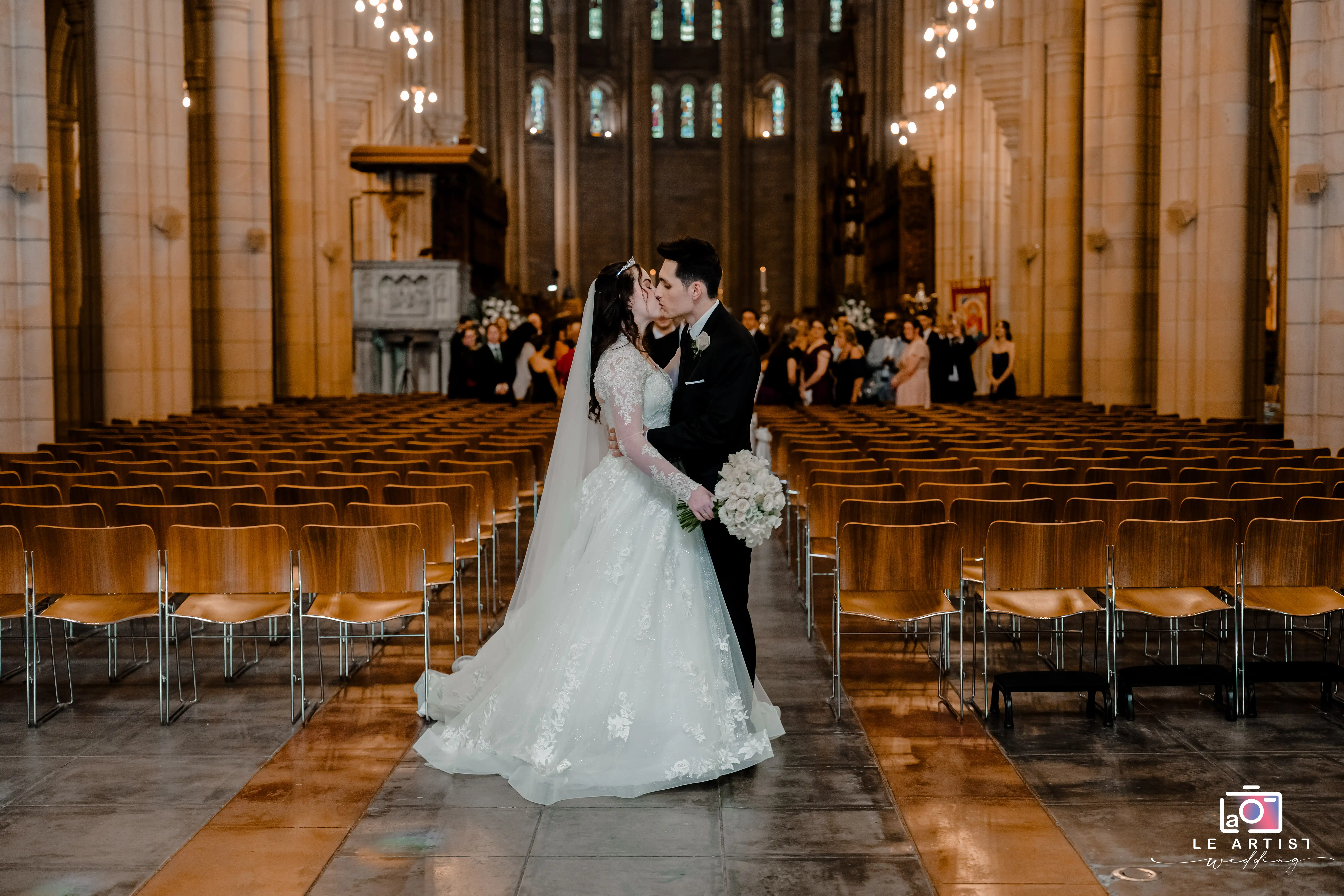 A romantic moment as the newlyweds share a kiss in an elegant church setting, captured beautifully with rows of chairs and guests in the background. Le Artist Wedding