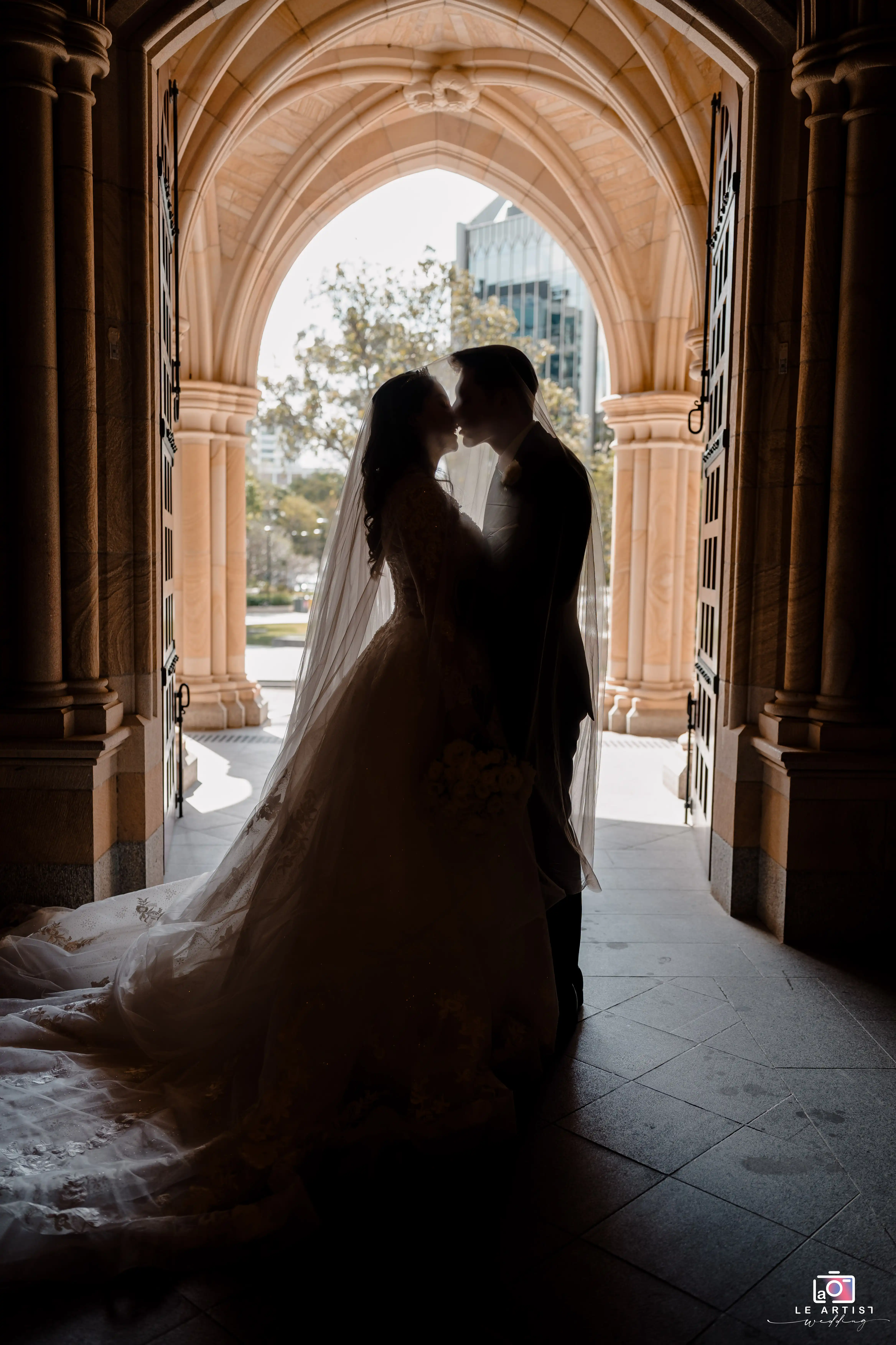 A romantic silhouette of the couple sharing a kiss under a grand archway, capturing the elegance and intimacy of the moment. Le Artist Wedding