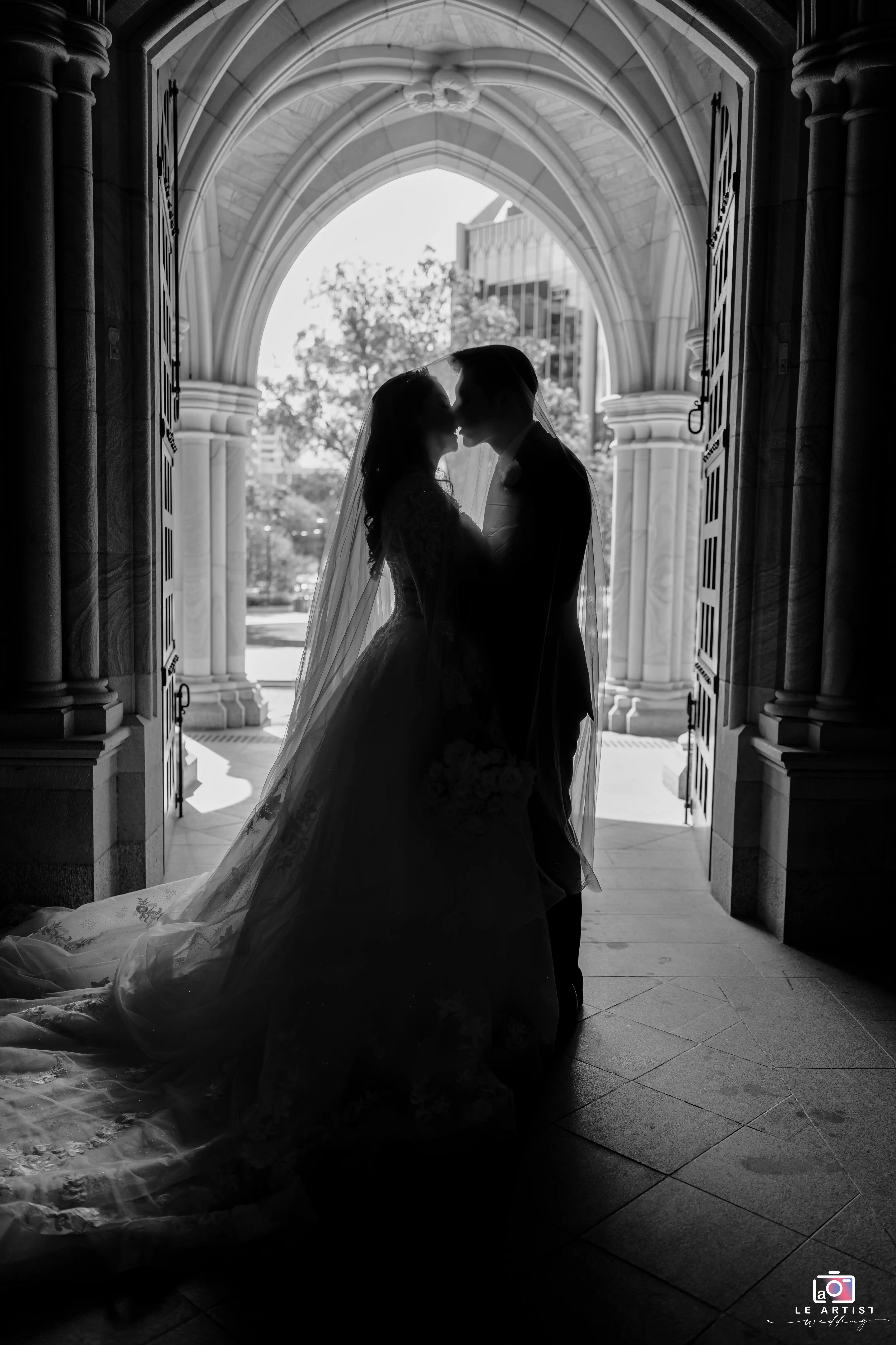 A romantic silhouette of a newlywed couple sharing a kiss in the arched doorway of a grand building. Le Artist Wedding