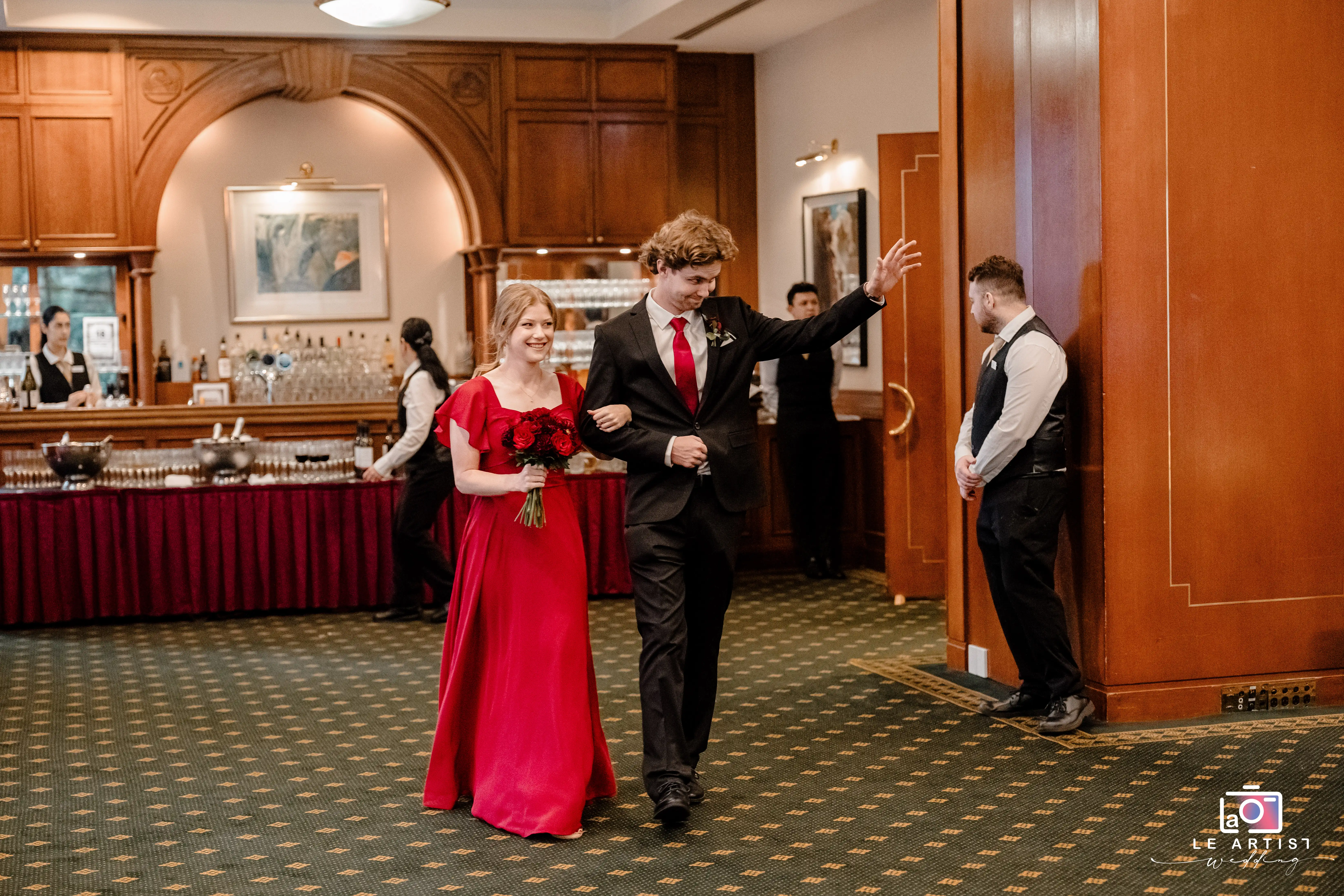 A couple makes a stylish entrance into the reception hall, radiating joy and confidence. The gentleman waves to the guests while the lady carries a bouquet of red roses, perfectly matching her elegant dress. Le Artist Wedding