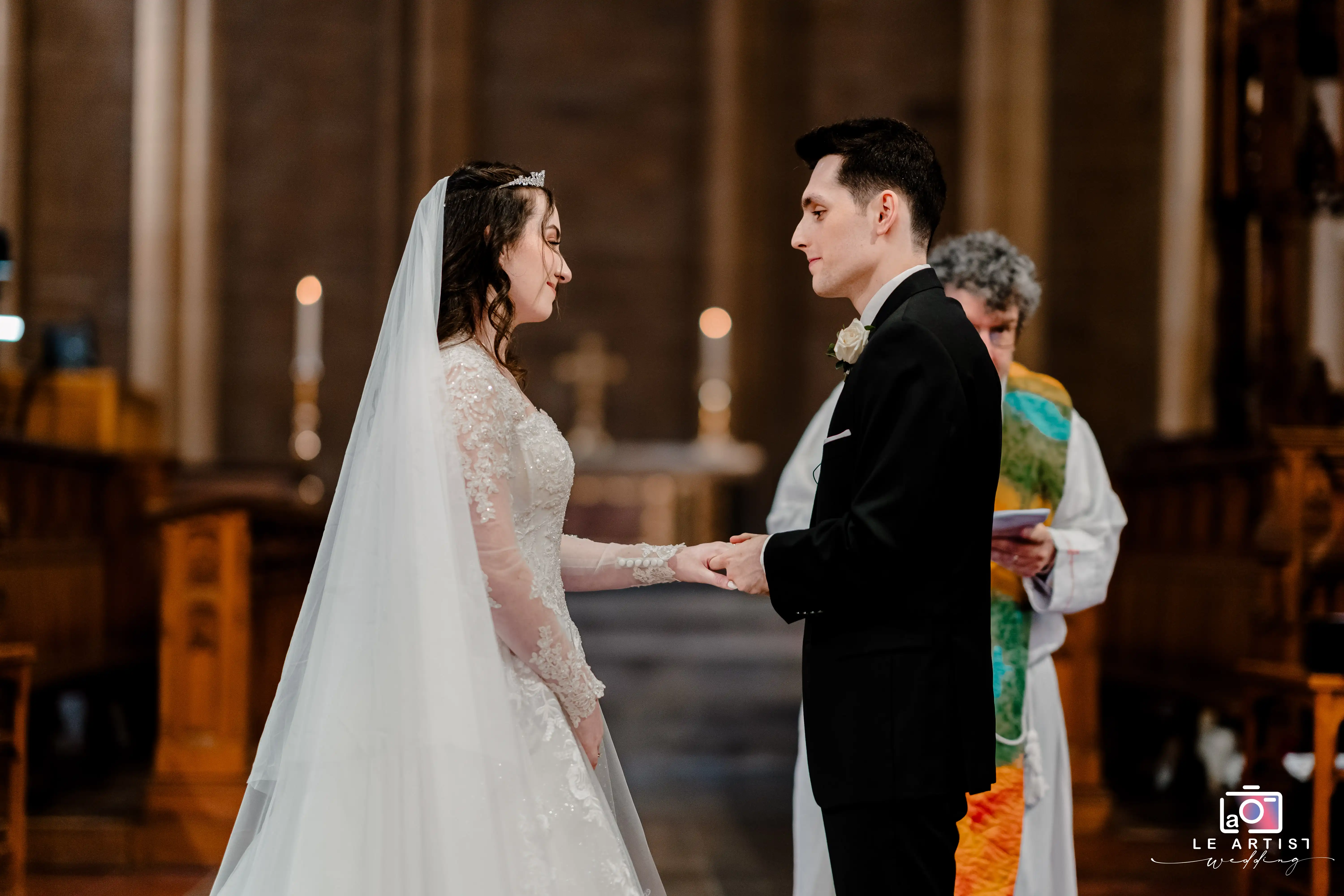 A heartfelt moment as the bride and groom exchange vows in a beautiful church setting, holding hands and gazing into each other's eyes under the gentle light of the altar. Le Artist Wedding