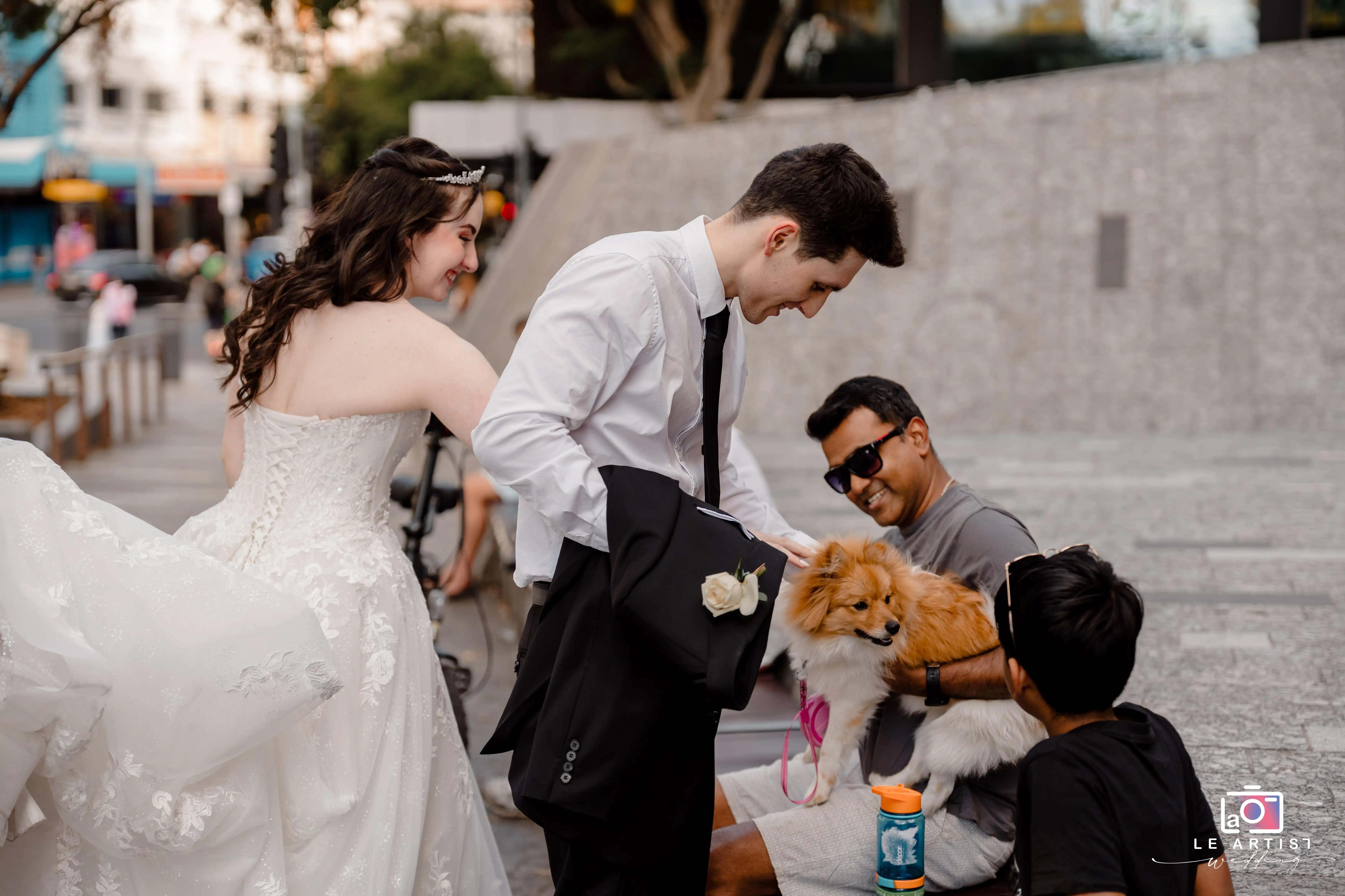 A joyful bride and groom share a light-hearted moment with passersby and a cute dog during their wedding day photo shoot. Le Artist Wedding