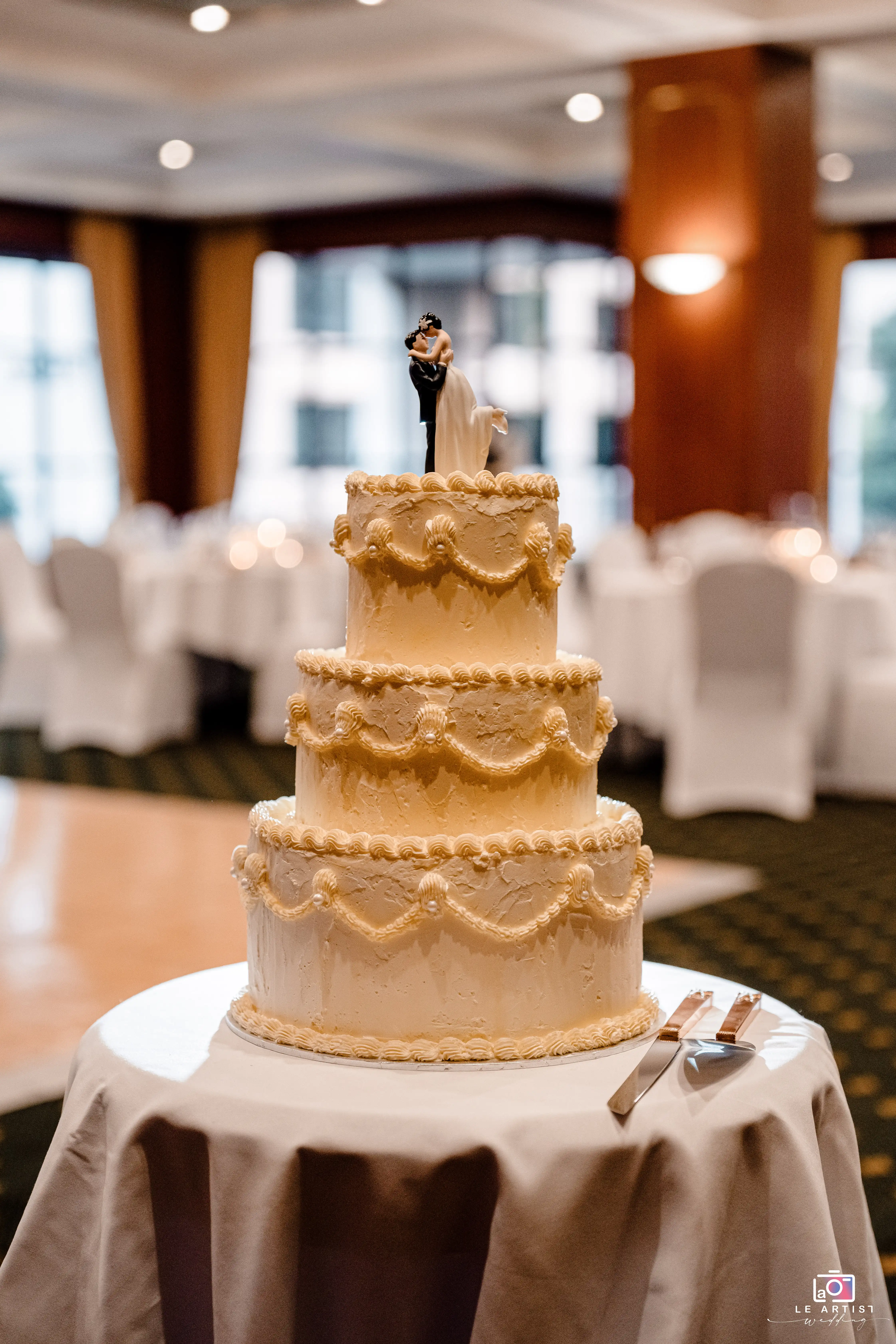 A beautifully decorated three-tiered wedding cake with a romantic couple topper is displayed in the reception area, ready for the cake-cutting ceremony. Le Artist Wedding