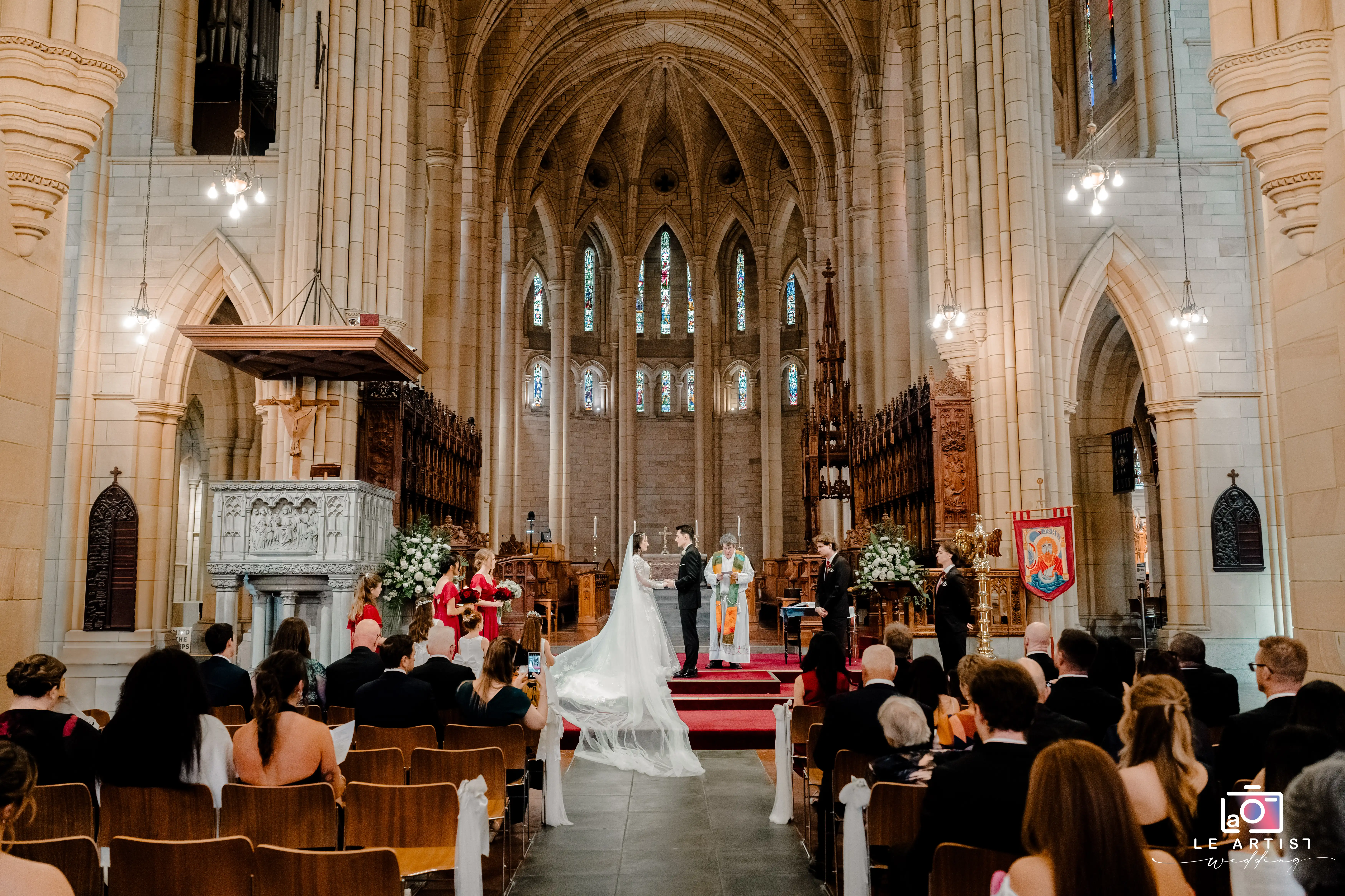 A captivating view of a couple exchanging vows in a grand cathedral, surrounded by friends and family, under magnificent arches and stained glass windows. Le Artist Wedding