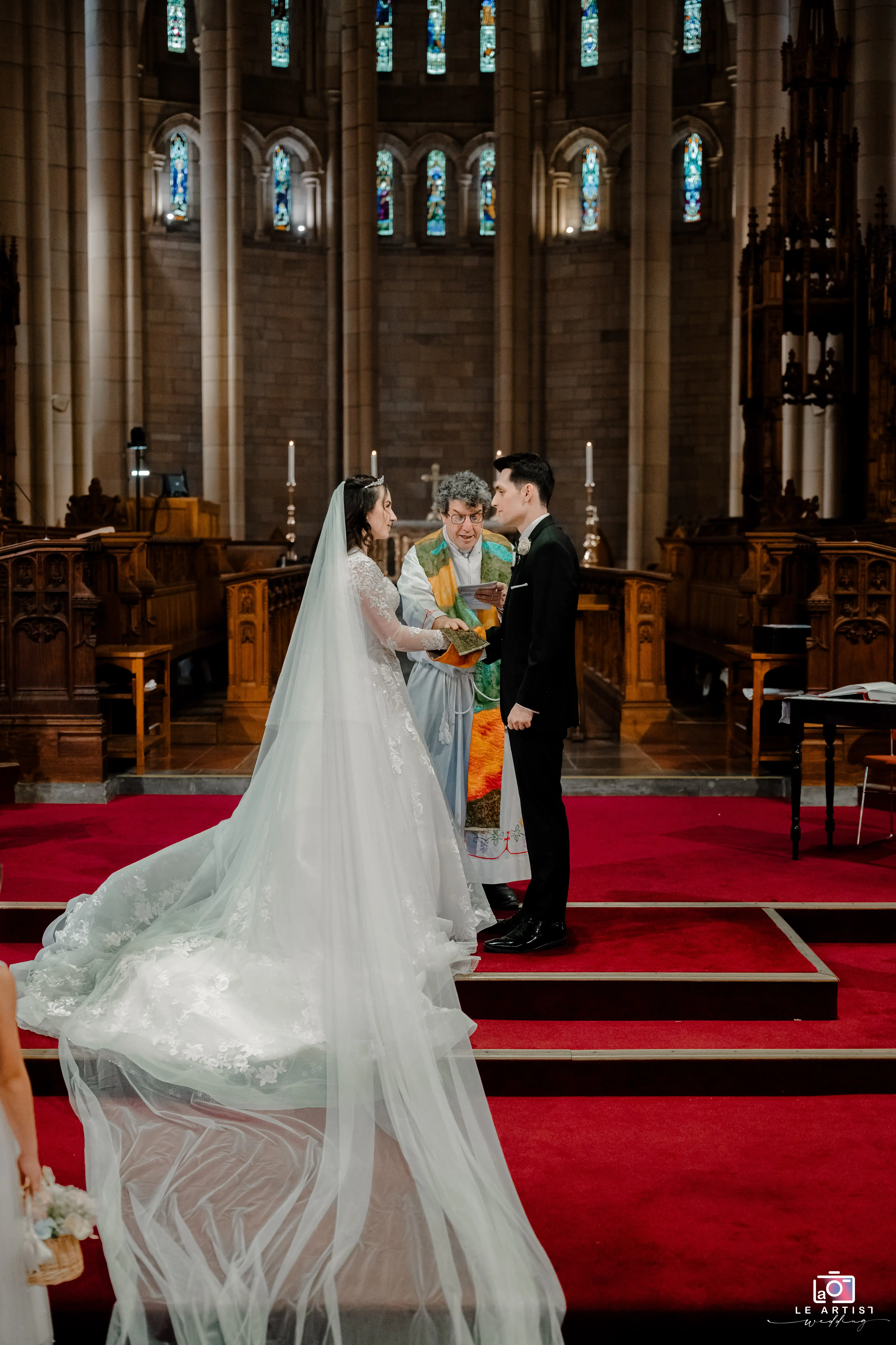 The couple stands before the officiant in a grand church setting, holding hands as they take their vows beneath stunning stained glass windows. Le Artist Wedding