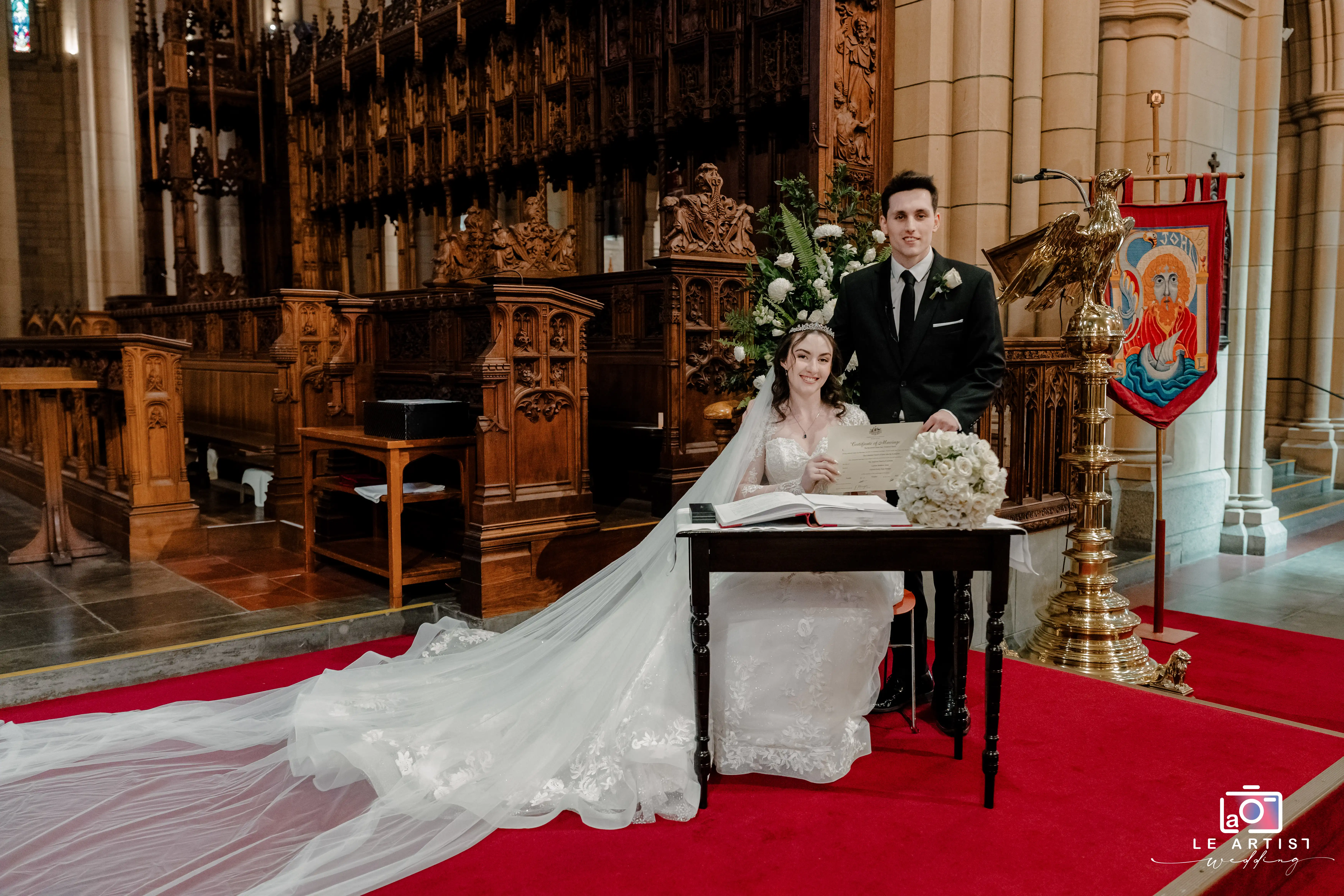 The newlyweds proudly display their marriage certificate inside a beautifully adorned church, surrounded by intricate wooden architecture and floral arrangements. Le Artist Wedding