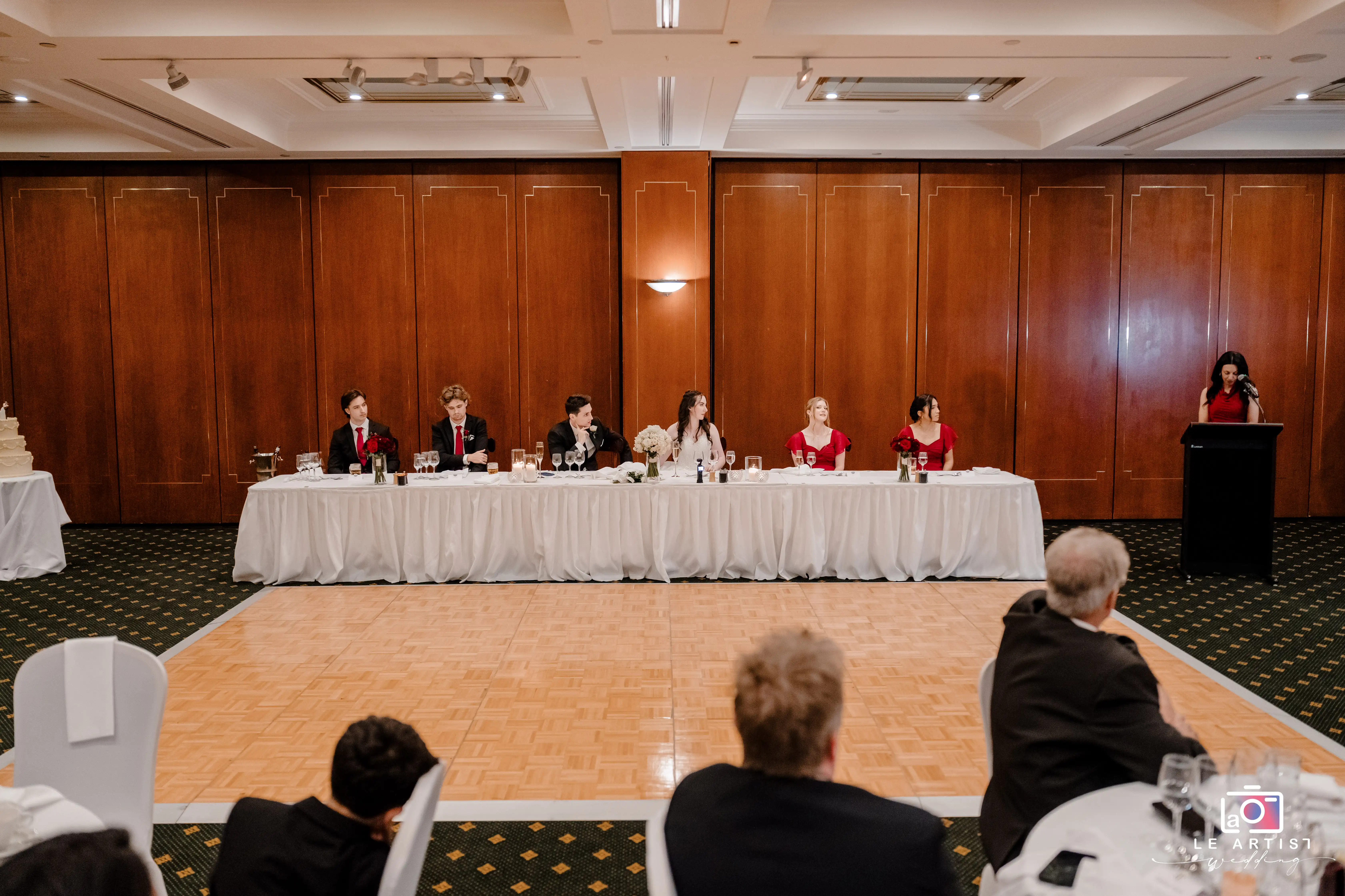 The wedding head table is elegantly set, surrounded by attendants in red and black attire. A speaker addresses the guests from the podium, adding a formal touch to this joyous occasion. Le Artist Wedding
