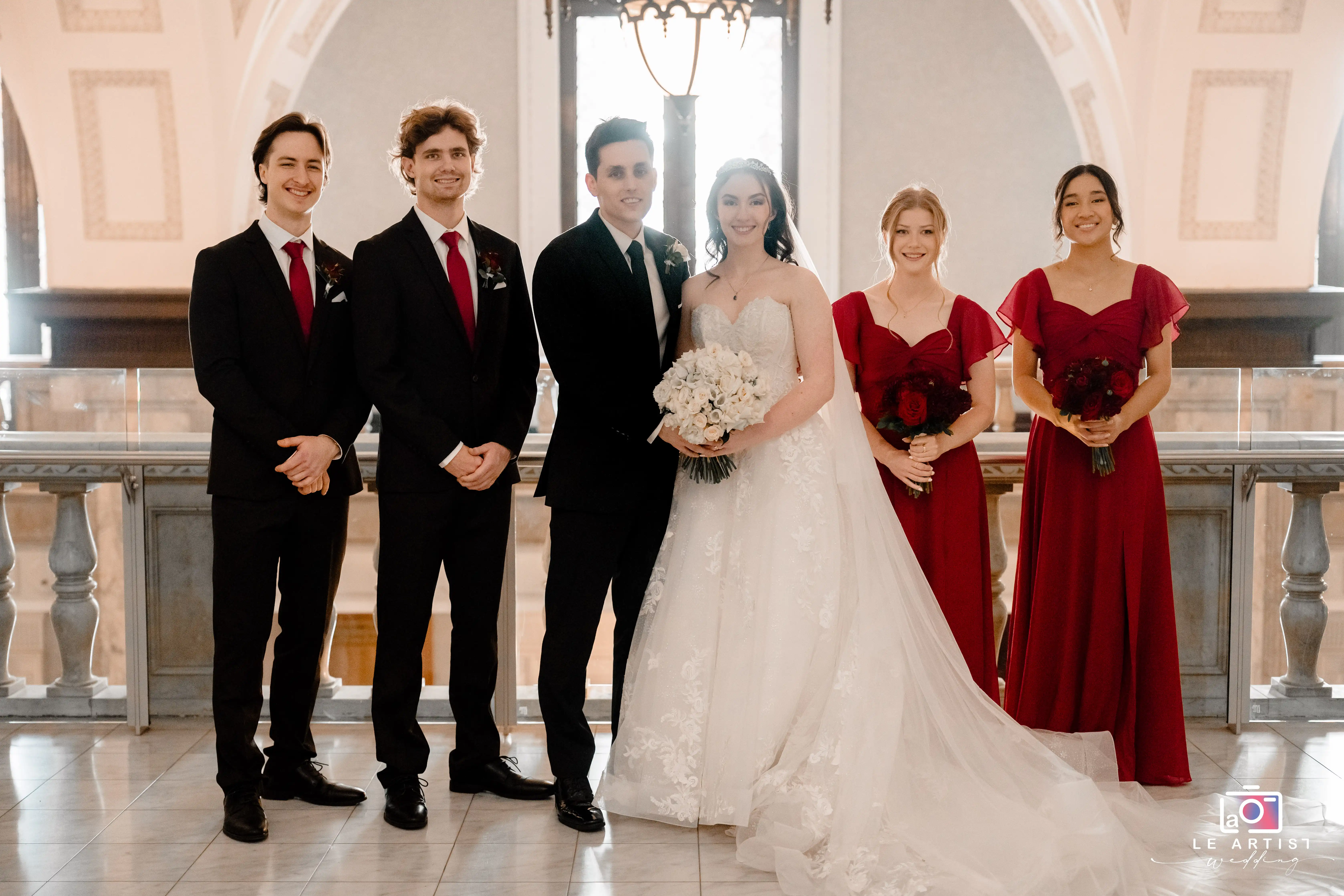 A joyful wedding party poses with the bride and groom, elegantly dressed in coordinating attire, set against a beautifully lit venue. Le Artist Wedding