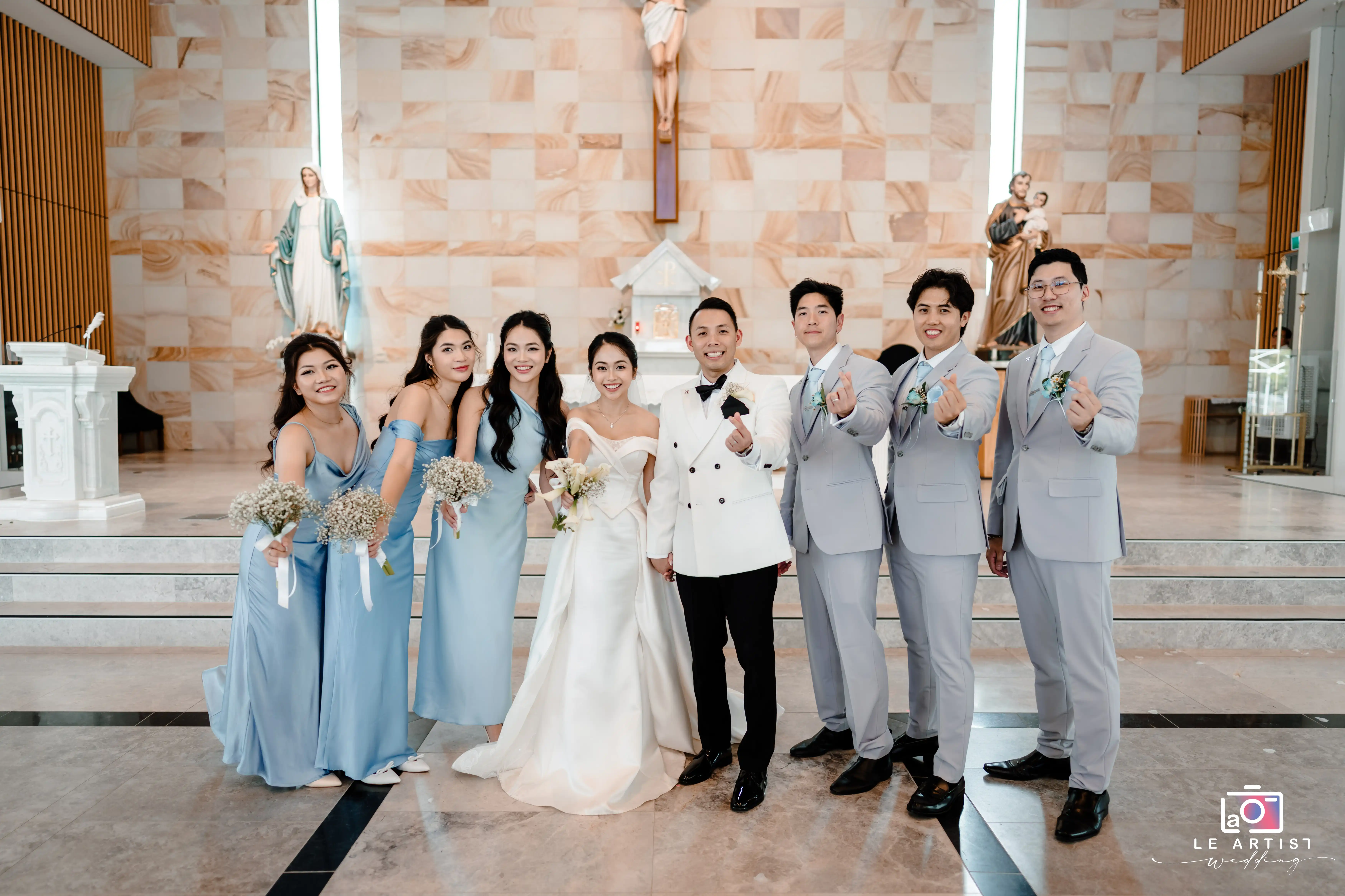 The newlyweds stand joyfully surrounded by their bridal party, showcasing happy and celebratory gestures in a beautifully adorned church setting. Le Artist Wedding