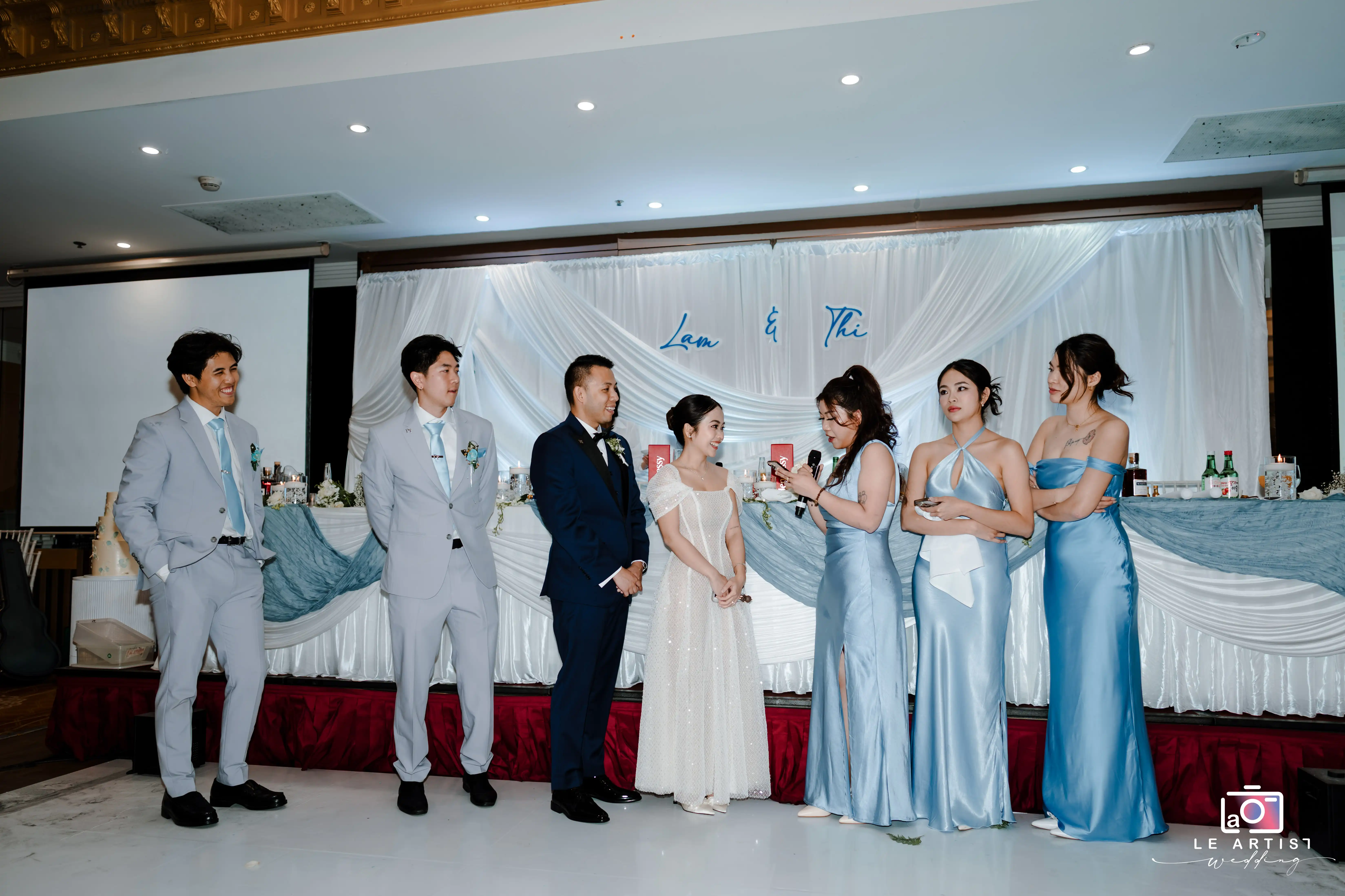 The bridal party stands elegantly on stage during a speech, with the bride and groom sharing a joyful moment amidst the beautifully decorated backdrop. Le Artist Wedding