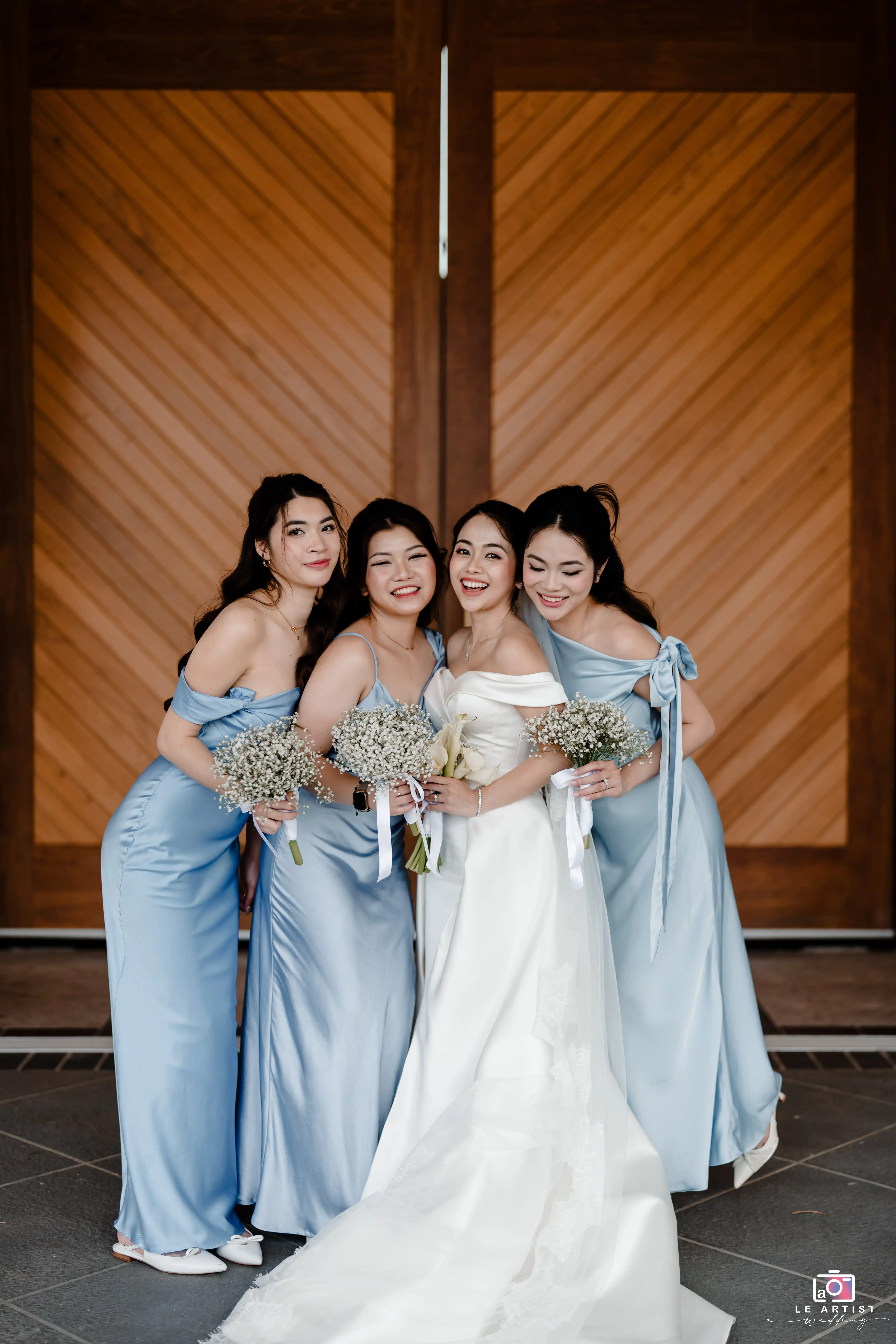 The bride and her bridesmaids pose together, radiating joy and elegance in their coordinated attire. Their bouquets add a charming touch to the group portrait. Le Artist Wedding