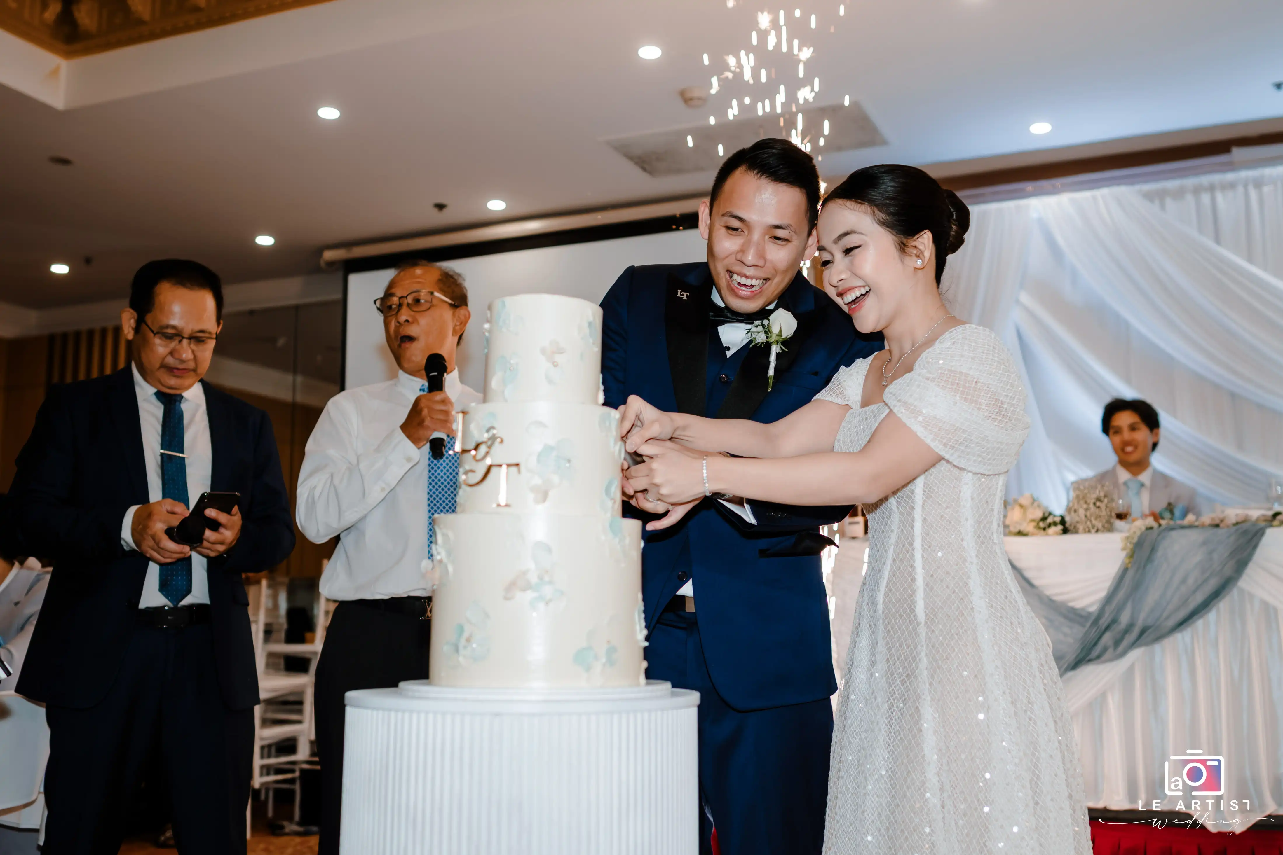 The bride and groom share a joyful moment as they cut their elegant multi-tiered wedding cake, surrounded by family and sparklers. Le Artist Wedding