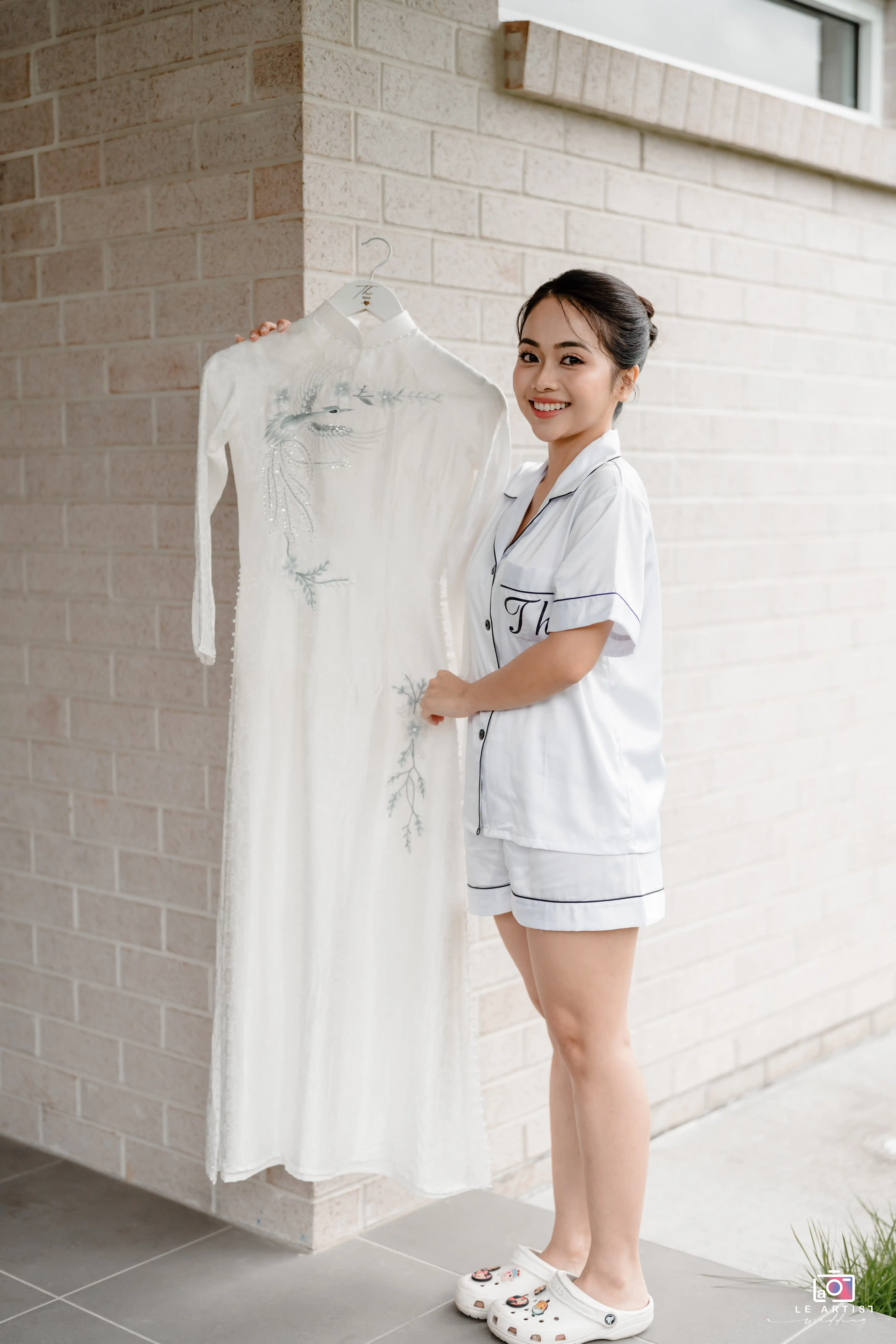 A bride joyfully admires her elegant white wedding dress embroidered with delicate patterns as she prepares for the special day. Le Artist Wedding