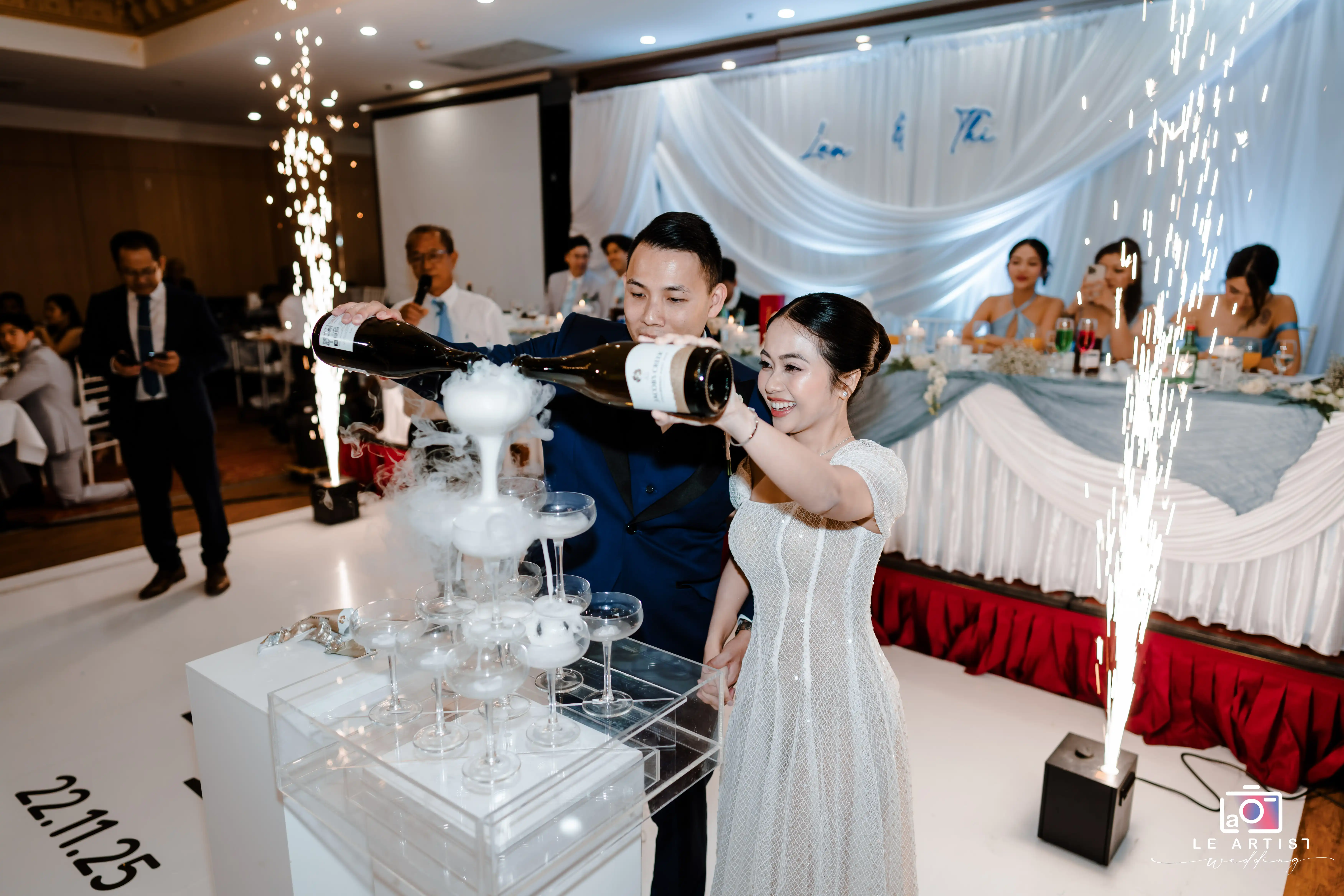 The couple joyfully pours champagne into a tower of glasses amidst sparklers at their wedding reception. Le Artist Wedding