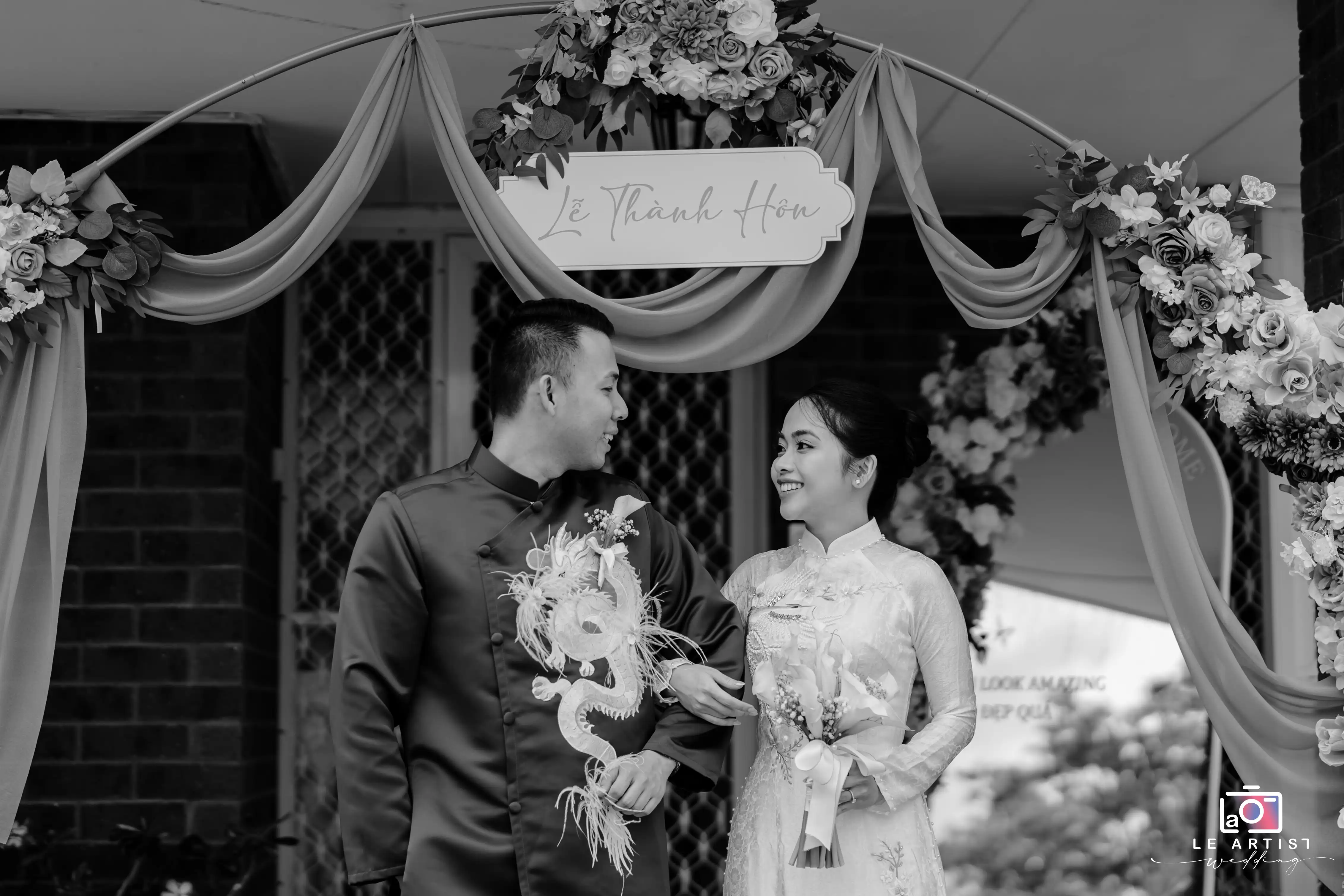 A couple shares a joyful moment under a beautifully decorated floral arch, capturing the essence of their celebration. Le Artist Wedding