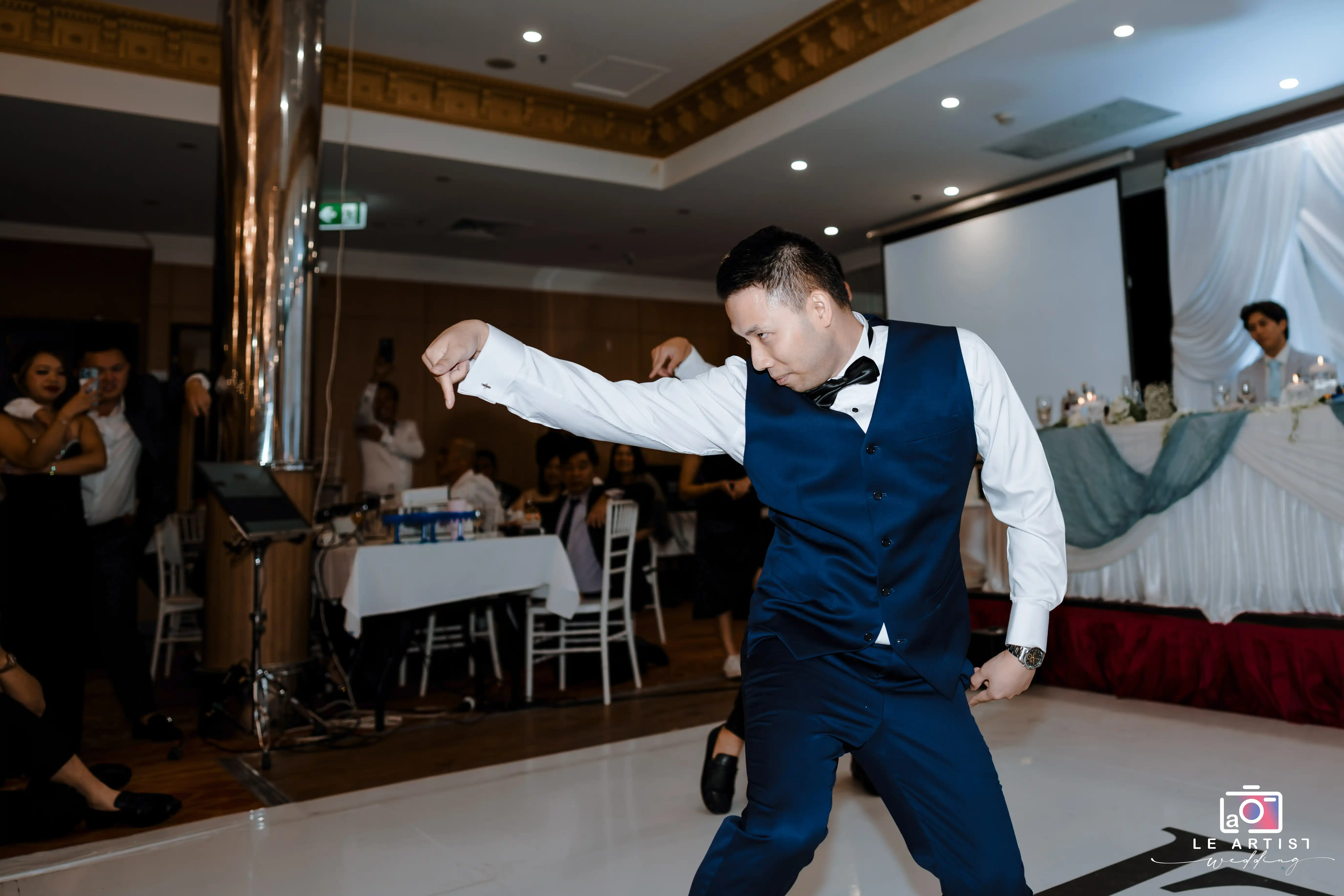 A lively wedding moment captures a man in a blue suit showcasing his dance moves on the dance floor, while guests watch and take photos. Le Artist Wedding
