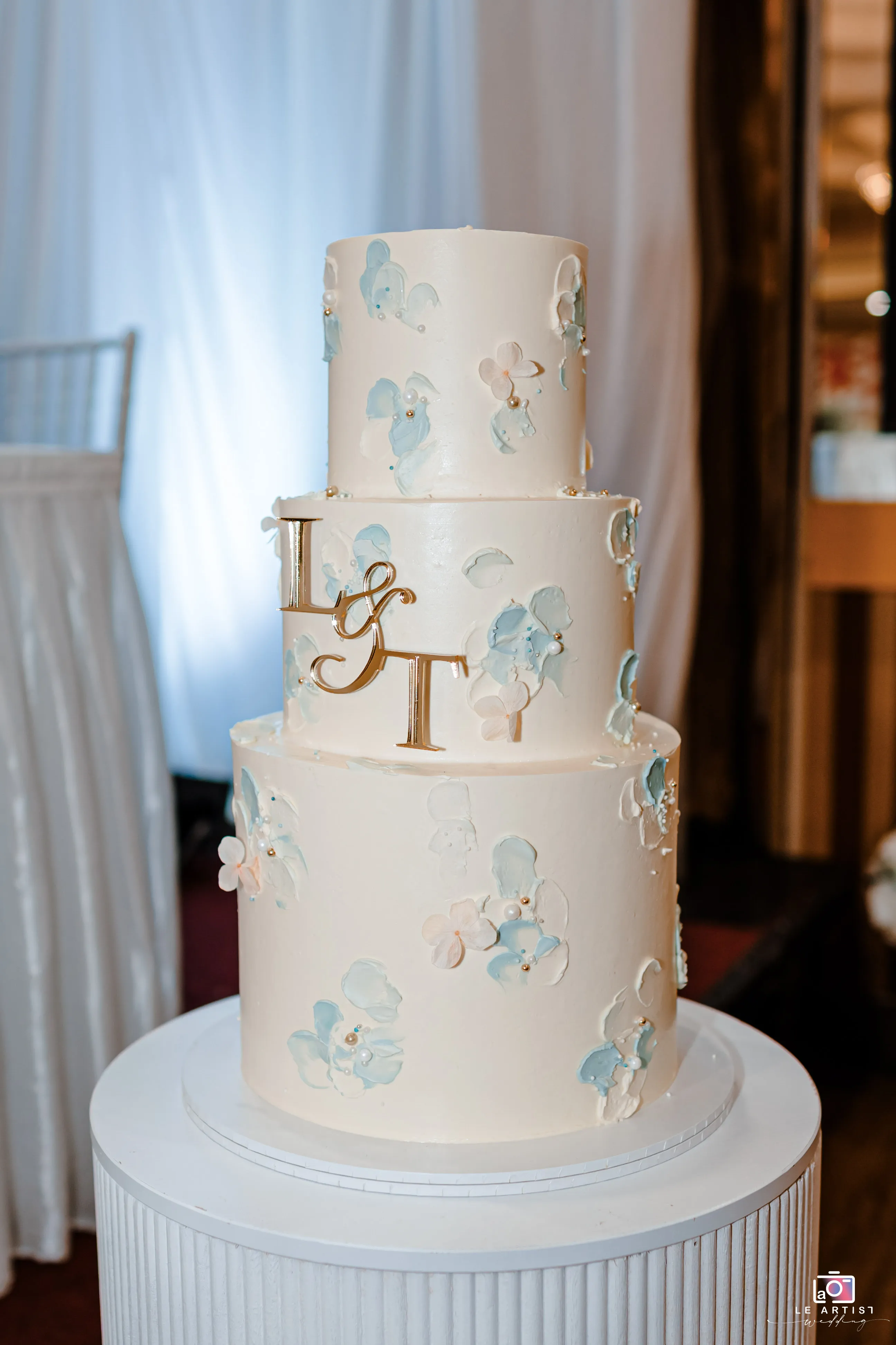 A beautifully decorated three-tiered wedding cake adorned with pastel blue and white floral designs, topped with a personalized monogram. Le Artist Wedding