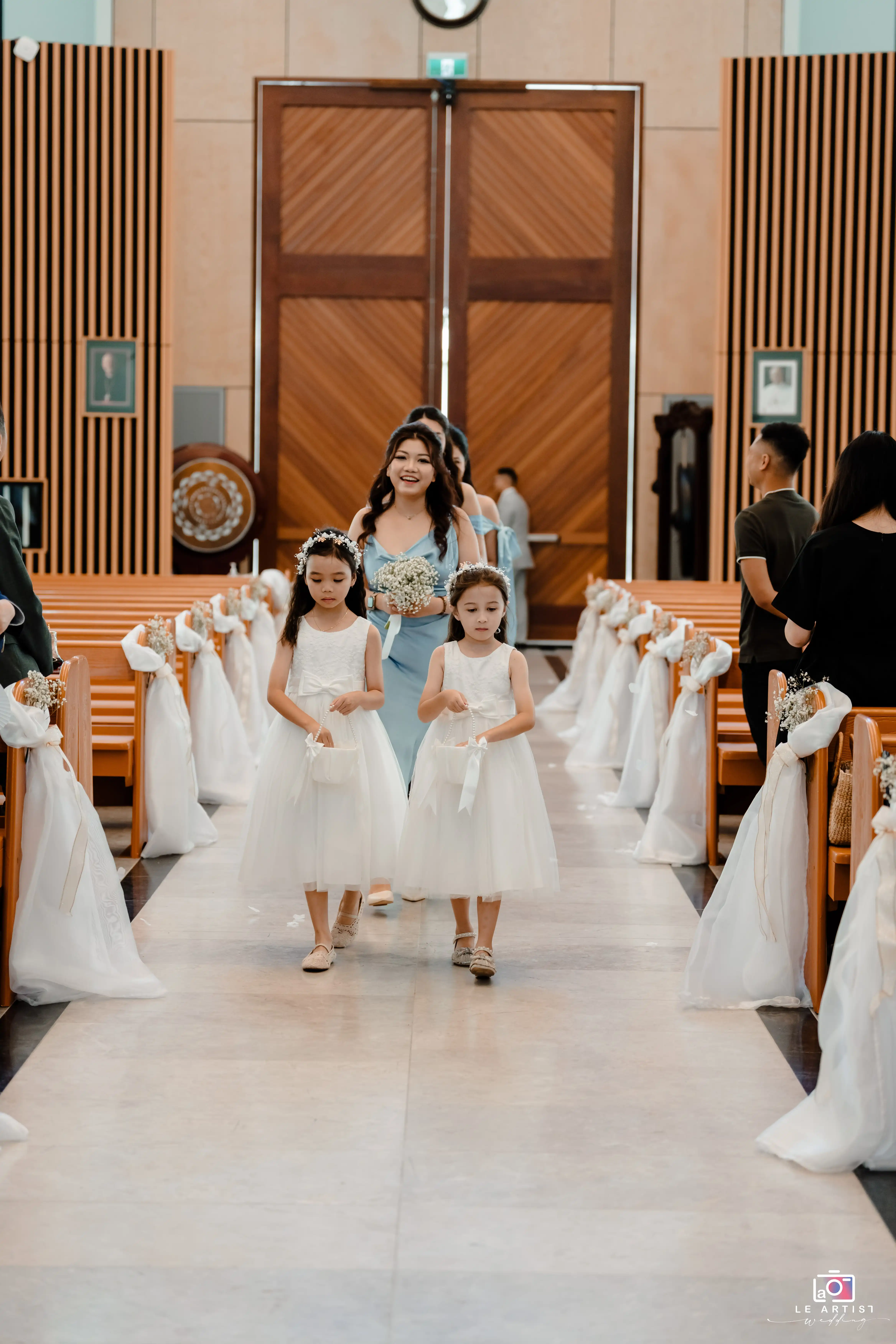 Flower girls leading the wedding procession down the aisle, holding small baskets, followed by bridesmaids in pastel dresses. Le Artist Wedding