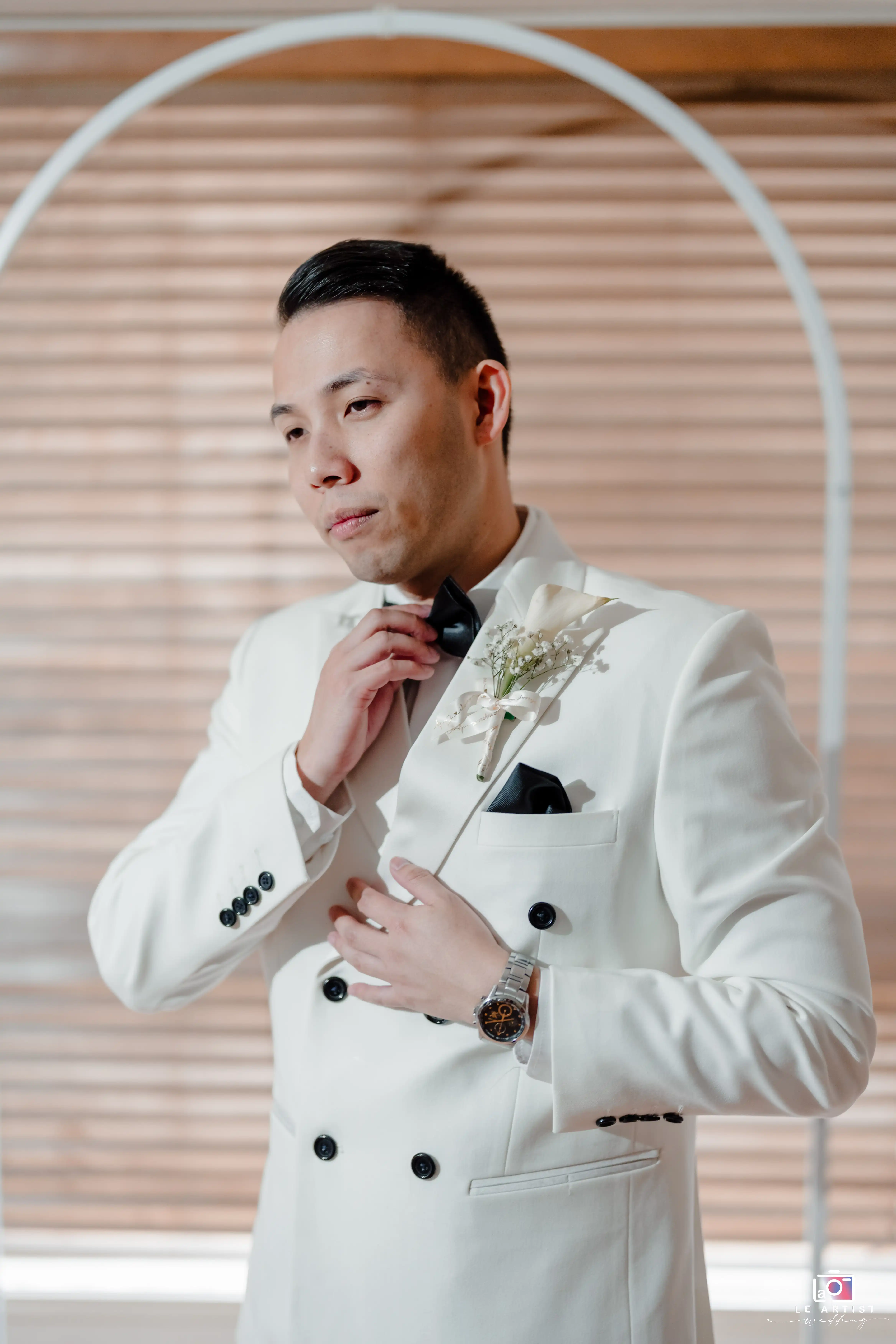 The groom adjusts his bow tie, looking confident in his elegant white suit with a touch of floral detail. Le Artist Wedding