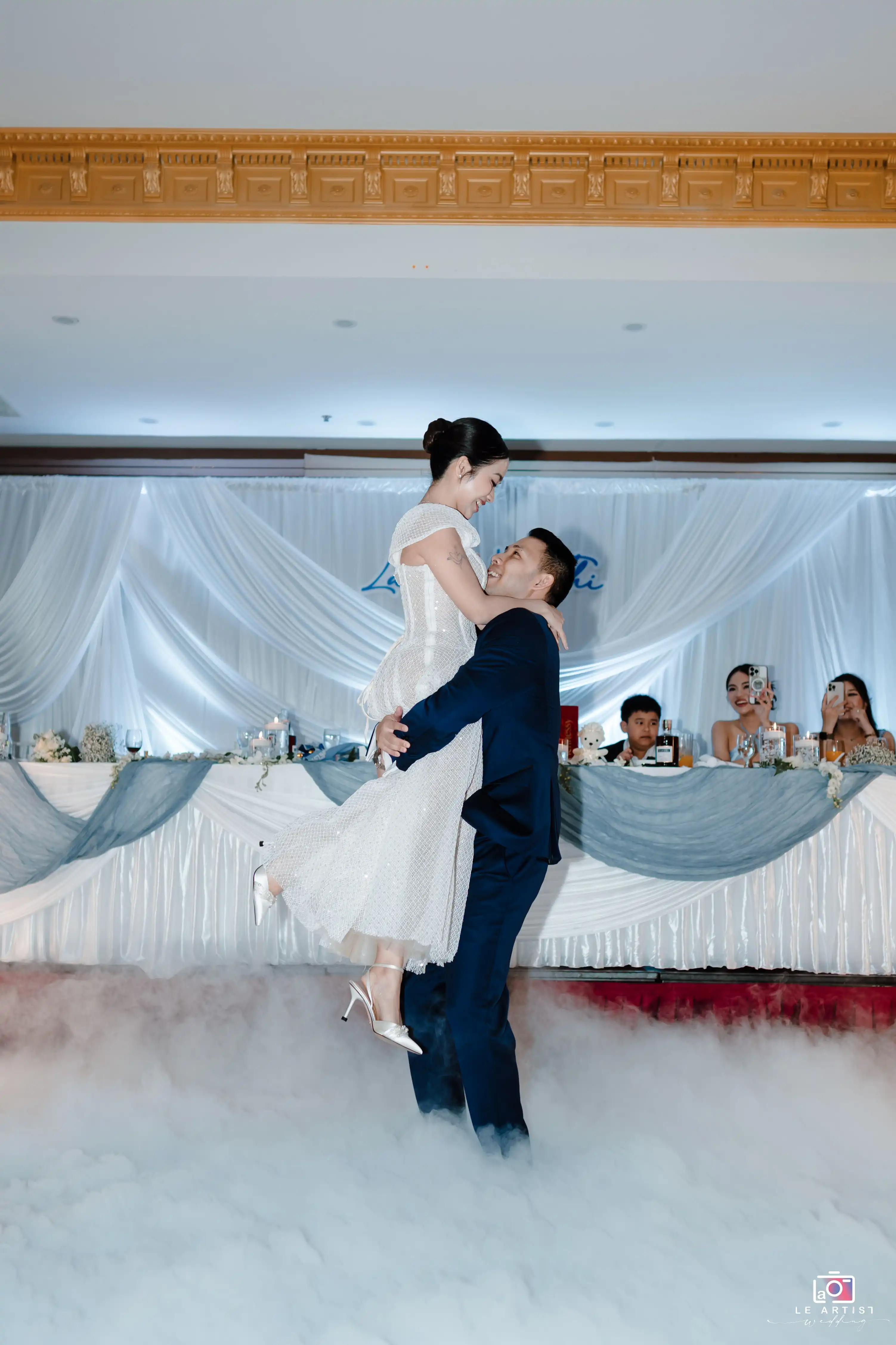 The couple shares a joyful moment as the groom lifts the bride during their dance, surrounded by a dreamy mist on the dance floor. Le Artist Wedding