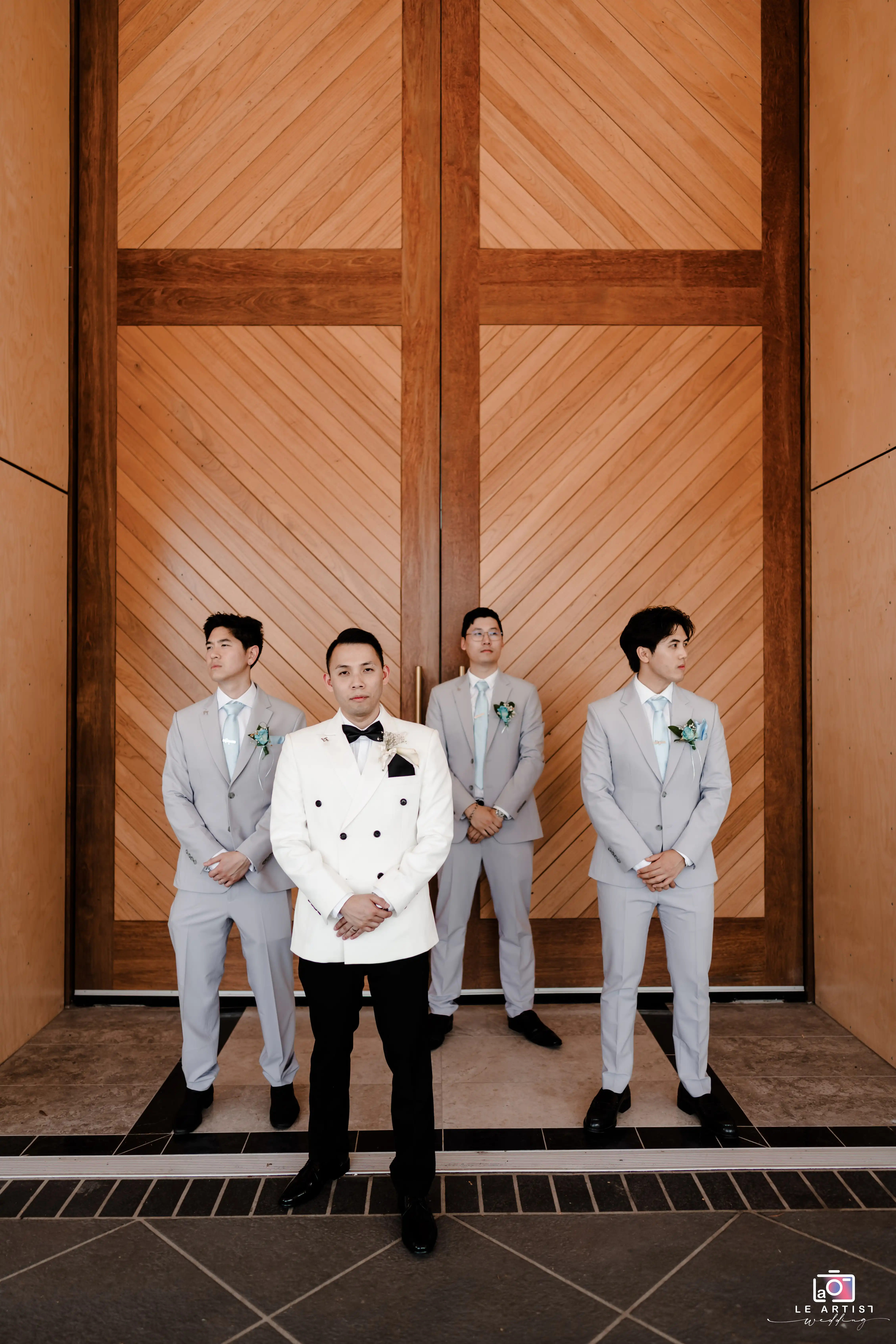 The groom stands confidently with his groomsmen in front of an elegant wooden backdrop, showcasing their style and camaraderie. Le Artist Wedding